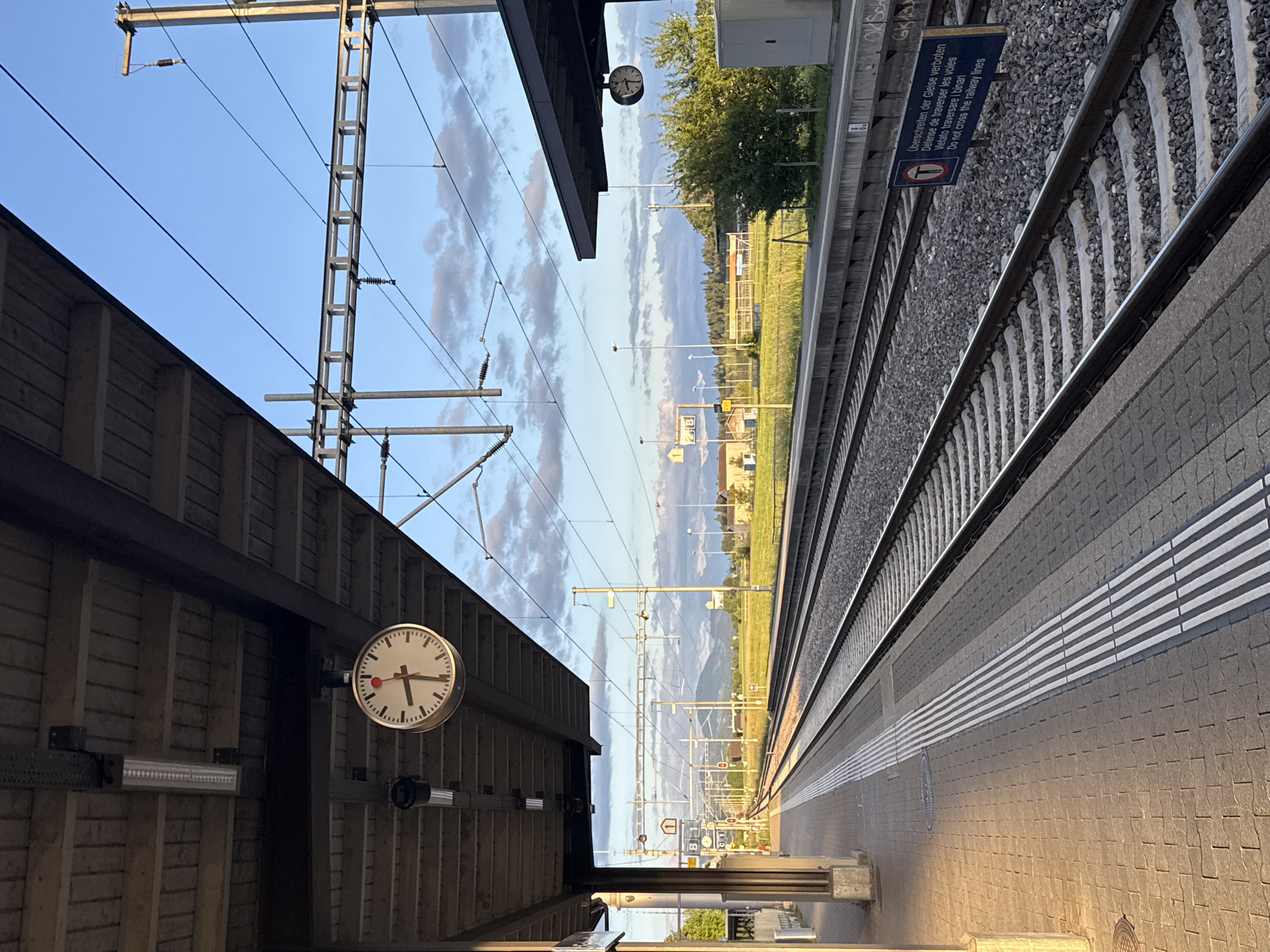 This image, correctly oriented, captures a serene late afternoon or early evening scene at a train station platform in Fehraltorf, Switzerland. The platform, paved with interlocking tiles and featuring a white striped safety line, occupies the right side of the frame, bathed in warm, golden sunlight. Two sets of railway tracks, complete with sleepers and gravel ballast, run parallel to the platform.
On the left, a station canopy with a wooden slatted wall is in partial shadow, contributing to the sense of time passing. A prominent classic Swiss railway station clock, characterized by its red second hand, is mounted beneath the canopy, indicating the time is around 4:50 PM or 5:00 PM. No people are present, emphasizing the quietness of the station.
Above the tracks, an extensive overhead catenary system of metal poles and wires is visible against a clear blue sky with scattered, wispy clouds. In the distance, beyond the tracks, the landscape features green fields, scattered buildings, and a backdrop of distant hills or mountains.
A notable detail is a blue warning sign positioned next to the tracks, which displays text in multiple languages: "Überschreiten der Gleise verboten," "Défense de traverser les voies," "Vietato traversare i binari," and "Do not cross the railway lines," along with a pictogram of a crossed-out person, clearly prohibiting track crossing. A smaller yellow sign marked "B" can also be seen further down the tracks, likely denoting a platform section. The overall mood is peaceful and calm under a pleasant sky.