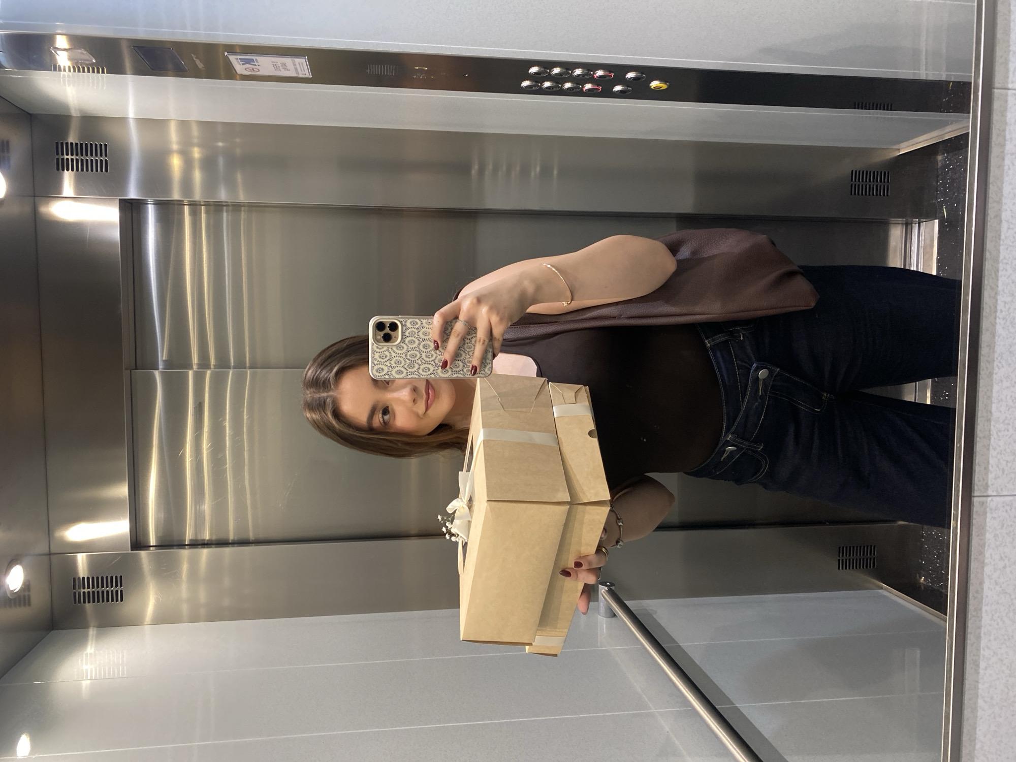 A young woman is taking a selfie in an elevator. She is holding a brown cardboard box with a white ribbon and a small bouquet of white flowers attached. The elevator has a mirrored interior, revealing a stainless steel wall with buttons and indicator lights at the top. The woman is wearing dark jeans and a brown top, with a brown purse slung over her shoulder. Her phone has a patterned case. The location appears to be an indoor setting, possibly a commercial or residential building, in the city of Unknown, Unknown.