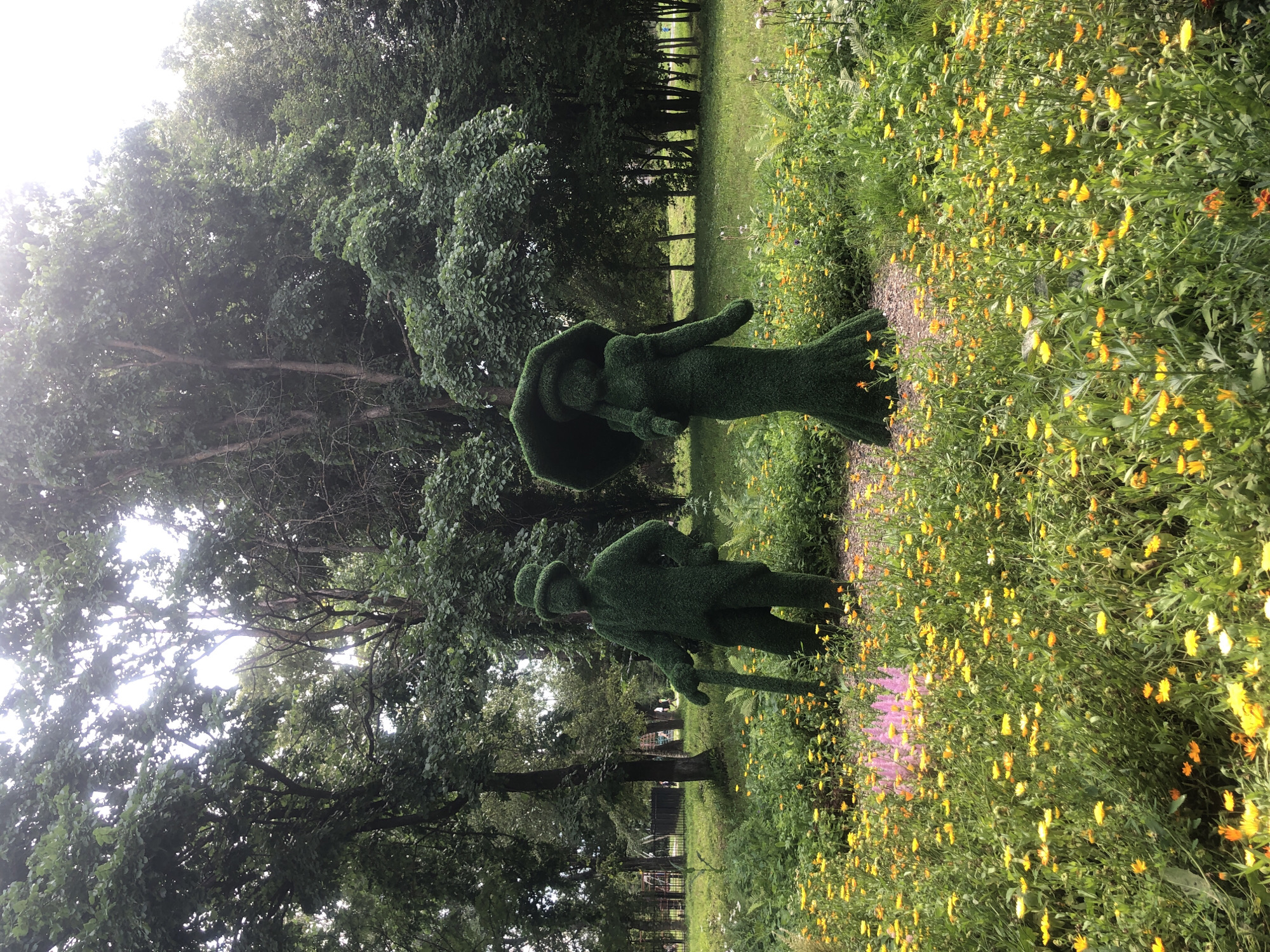 The image captures a vibrant park scene in Tutaev, Russia, featuring two topiary figures set amidst a colorful meadow of yellow and orange wildflowers. The figures, one resembling a man with a cane and hat, and the other a woman holding an umbrella, are sculpted from lush green vegetation, creating a whimsical and artistic display. Tall trees form a verdant backdrop, their foliage dense and lush, suggesting a warm, summer day. The park appears well-maintained, with manicured grass areas and a pathway visible in the distance, hinting at a public space designed for recreation and aesthetic appreciation. The overall atmosphere is one of tranquility and natural beauty, enhanced by the charming topiary sculptures.