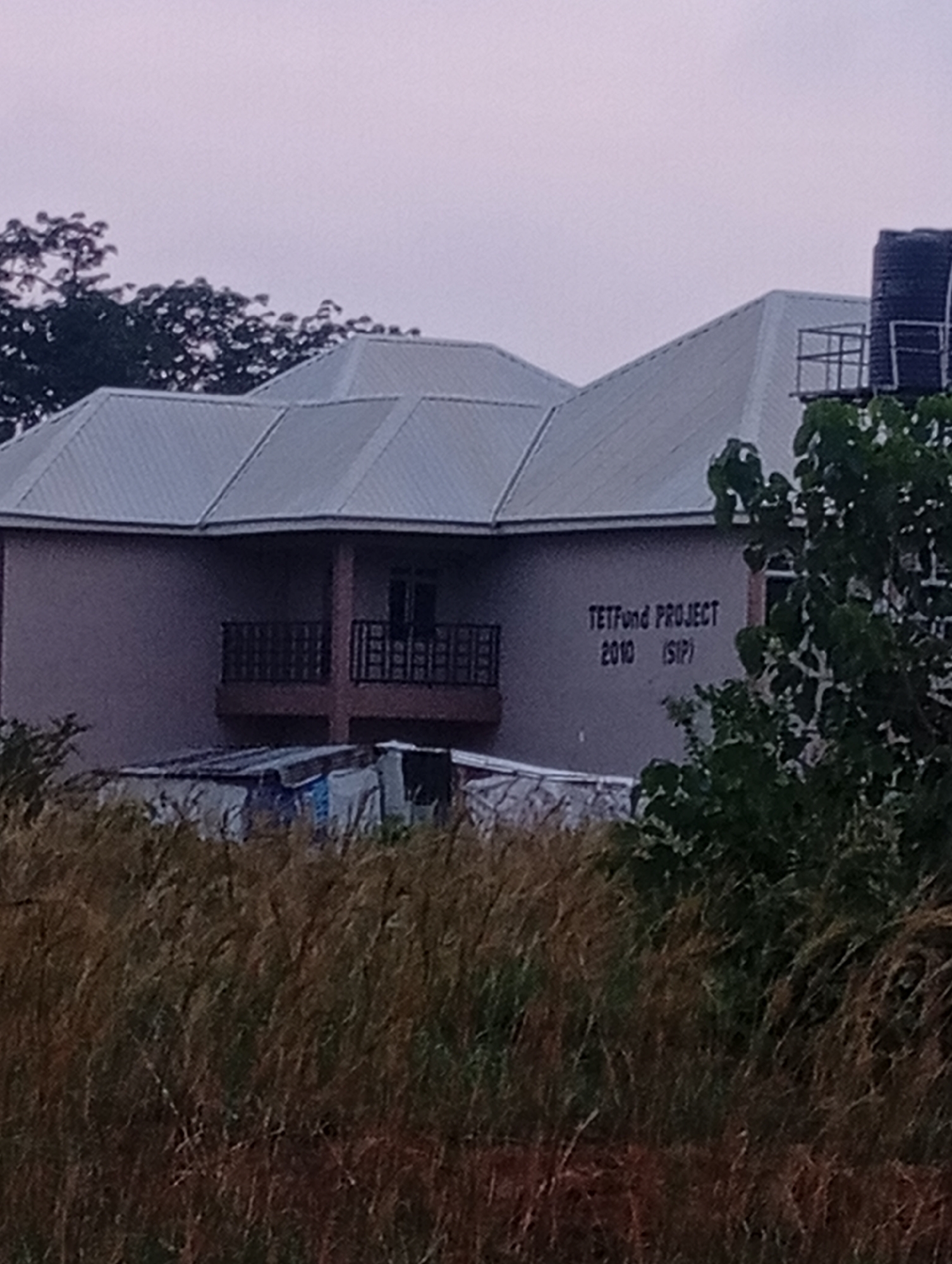 A medium shot captures a two-story building with a grey, pitched roof and beige walls. The building is situated in Du, Nigeria, amidst tall, dry grass in the foreground and trees in the background. On the upper story, a balcony with a black metal railing extends across the center. Text on the wall reads "TETFUnd PROJECT 2010 (SIP)". To the right of the building, a large, black water tank is visible on a raised platform. The sky is overcast and pale.