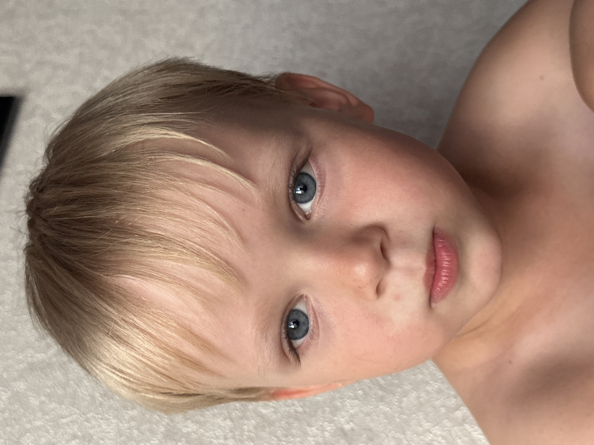 A close-up, eye-level, portrait photograph shows the face of a young Caucasian boy with fair skin, light blond hair, and blue eyes. He is looking directly at the camera with a neutral expression. The background is a textured, light-colored wall, possibly made of concrete or plaster. The boy's shoulders and upper chest are visible, and he appears to be shirtless. There are no other people or objects in the frame. The lighting suggests an indoor setting. The city of Energetik, Russia is not depicted in this image.