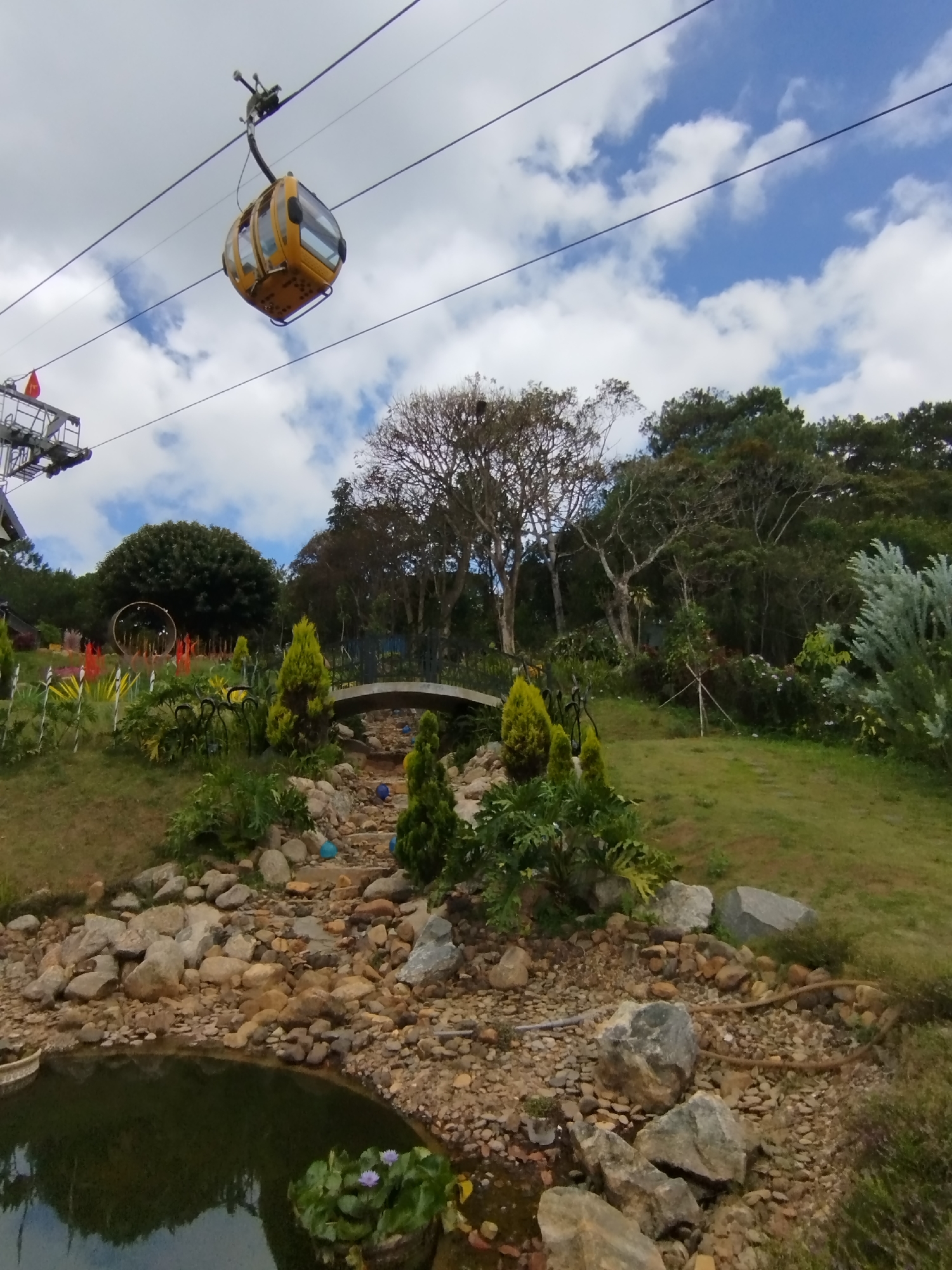 A landscape scene shows a yellow cable car traveling along overhead wires in Da Lat, Vietnam. The ground is a tiered garden leading to a still pond with floating lotus plants. A small, arched bridge crosses a dry streambed filled with rocks and blue decorative balls. Various trees and plantings populate the landscape, including ornamental trees. In the background, additional structures and foliage create a lush backdrop under a sky with scattered clouds.