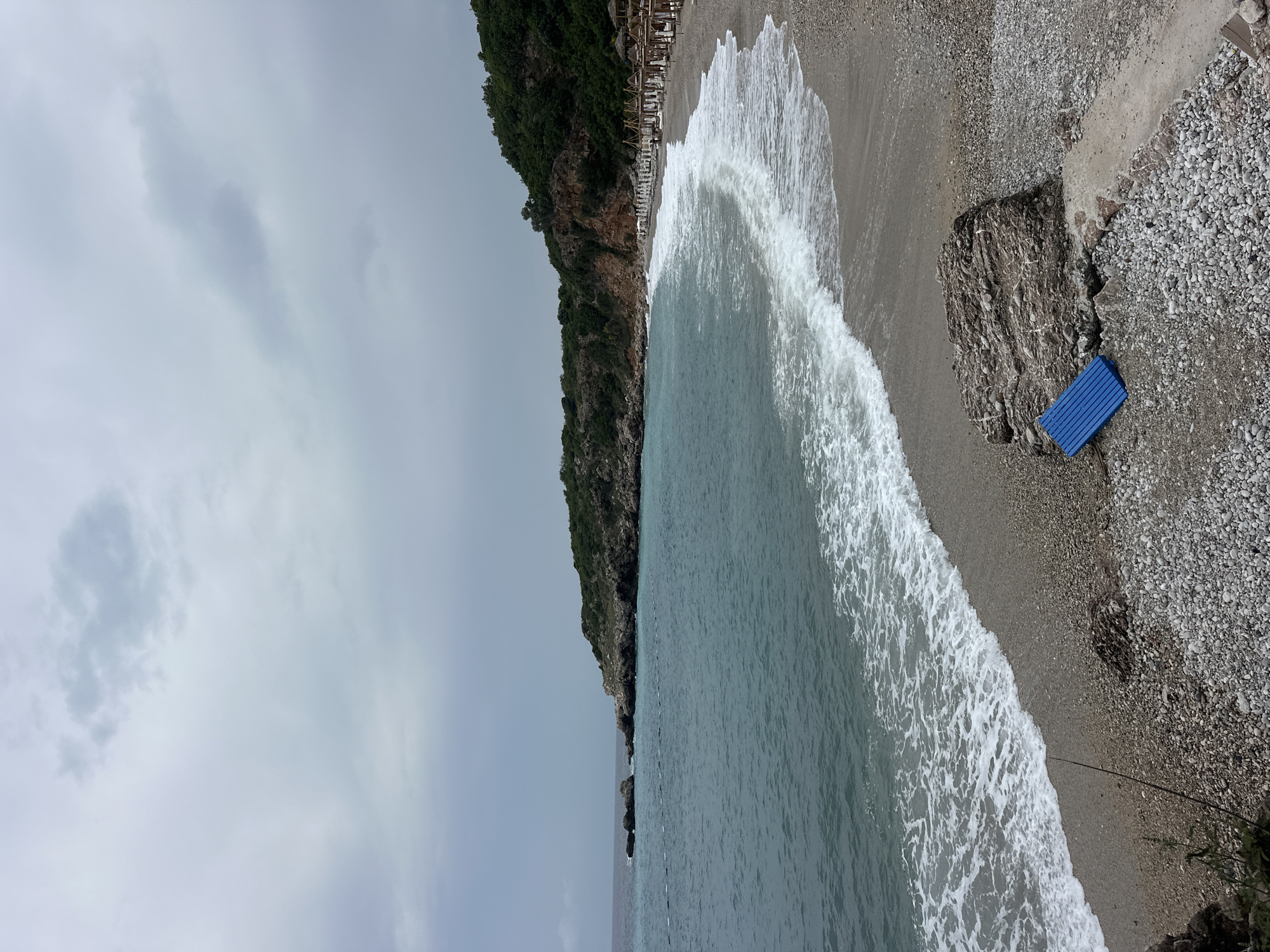 This media file presents a scenic coastal landscape at Drobnići, Montenegro, under an overcast sky. The image captures a wide view of a pebble beach bordered by the sea on one side and a steep, tree-covered cliff on the other.
The left half of the scene features the light blue-green waters of the sea, with white, foamy waves breaking gently onto the pebbled shore. A faint line of white buoys or markers extends from the left foreground into the water, suggesting a designated area.
The beach itself is composed of light-colored pebbles and gravel. Along the base of the dark green, vegetated cliff that backs the beach, numerous sun loungers with light-colored umbrellas are neatly arranged in rows, indicating an organized leisure area. Further to the right on the beach, a prominent, dark grey rock formation stands out, and next to it, a bright blue, rectangular object, possibly a mat or small board, lies on the pebbles.
The sky above is completely overcast with soft grey clouds, giving the entire scene a muted and tranquil atmosphere. The light suggests it is daytime, but the heavy cloud cover prevents direct sunlight. No people are visible in the image, contributing to a sense of peaceful solitude or suggesting an off-peak time. There is no visible text in the image.