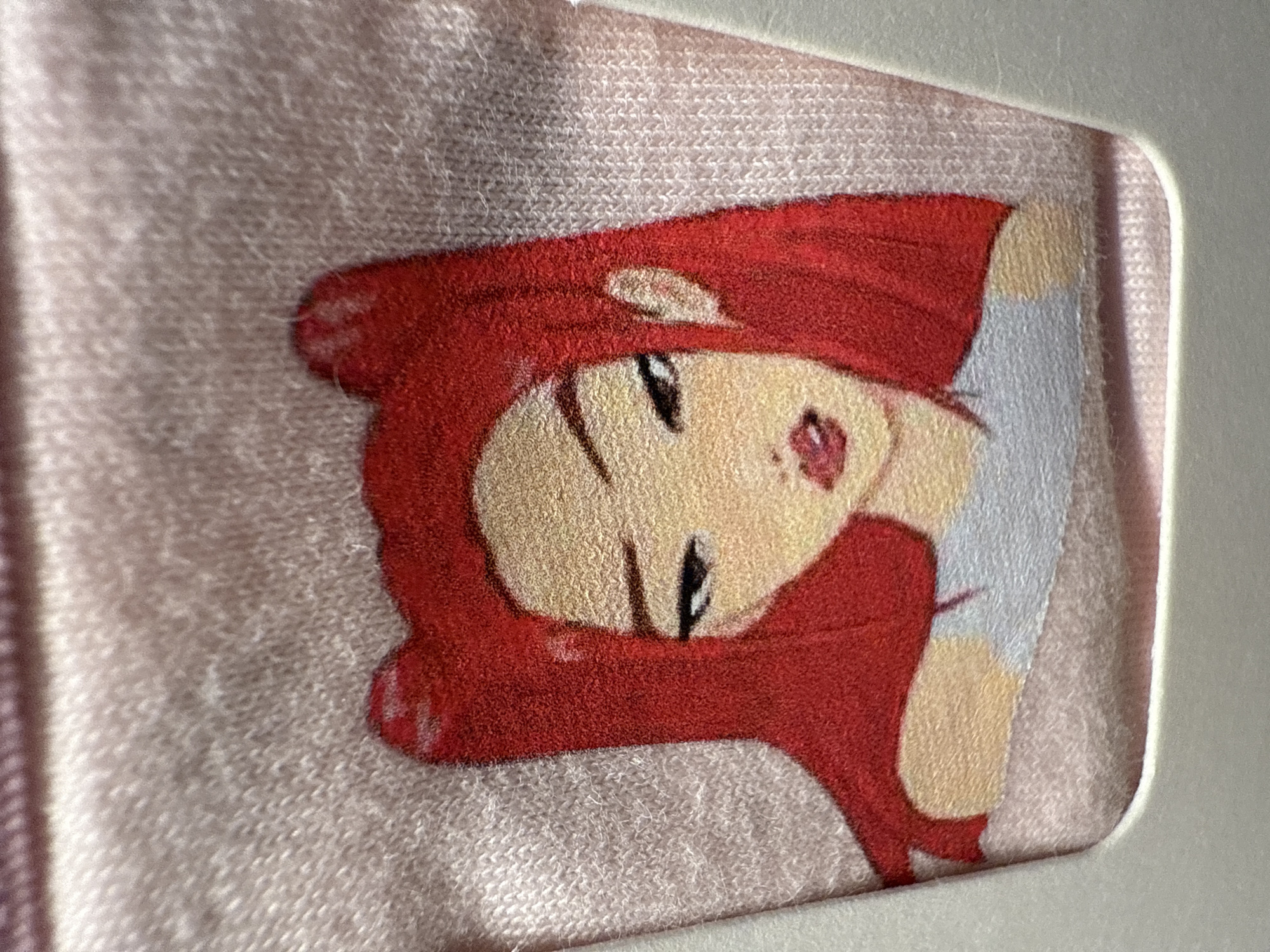 A close-up, slightly angled shot captures an anime-style female character depicted on a light pink, knitted or woven textile. The character features striking red hair that frames her fair-skinned face. Her eyes are closed, outlined in black with delicate lashes, and her mouth forms a gentle, contented smile. A small, heart-shaped red detail is visible near her left cheek. Parts of her clothing or accessories, rendered in white and red, are visible around her. The distinct, slightly fuzzy texture of the underlying fabric, possibly fleece or felt, is evident across the entire image, including within the printed areas of the character. A clean, white border with rounded corners is visible along the right and bottom edges, suggesting the textile is a patch, a label, or part of an item with a defined frame. No activity is taking place, and no text is visible. The image was taken in Miami, United States of America.