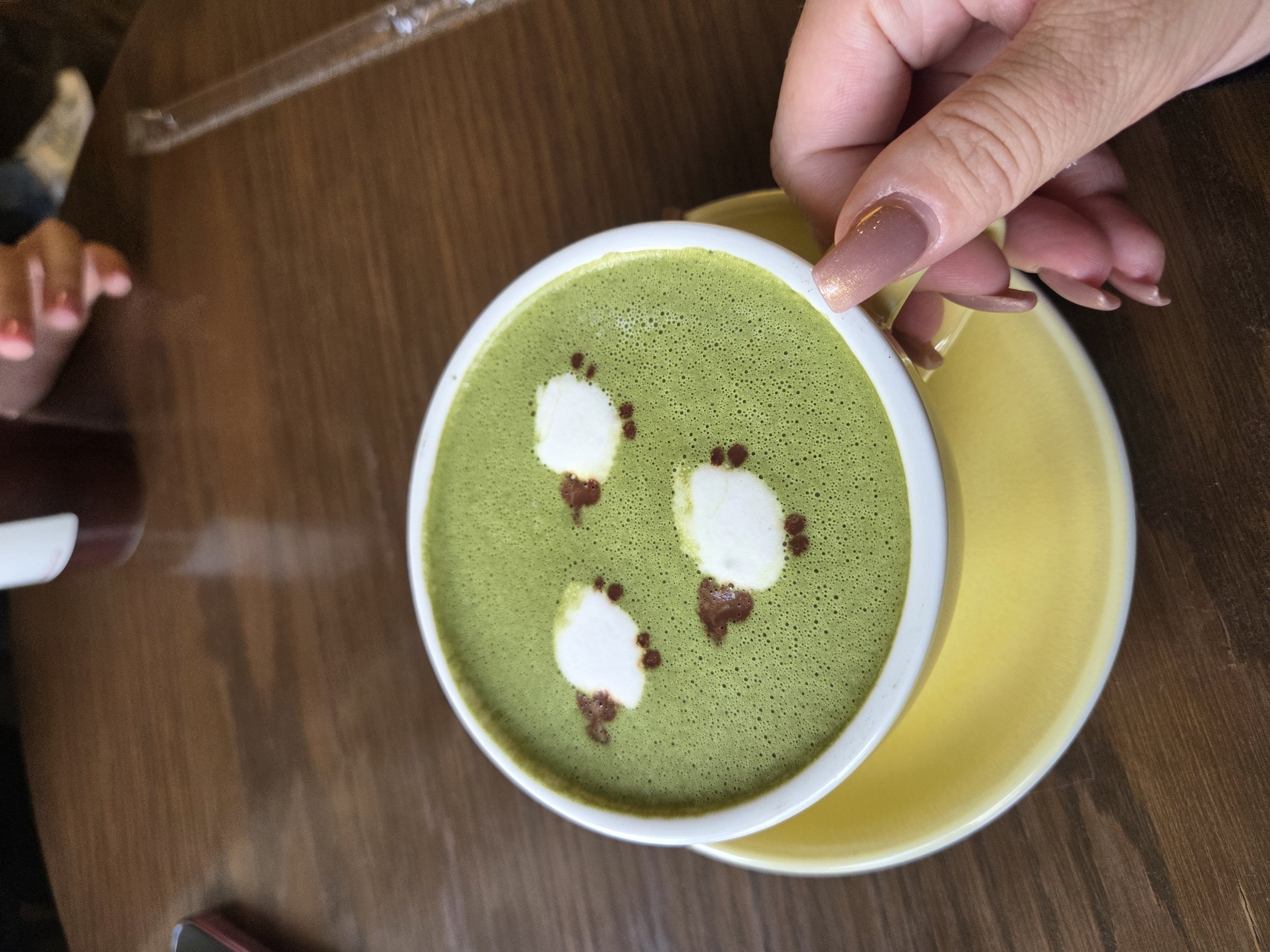 A close-up overhead shot shows a white mug with green matcha latte art resembling sheep, placed on a yellow saucer. The mug and saucer are on a dark wooden table. A person's hand with light pink nail polish is holding the mug from the side. In the background, parts of other objects are visible, including a dark container, a phone, and a clear plastic straw. The scene appears to be in a cafe or restaurant setting. The provided information that the location is in Minsk, Belarus is not visually verifiable from the image itself.