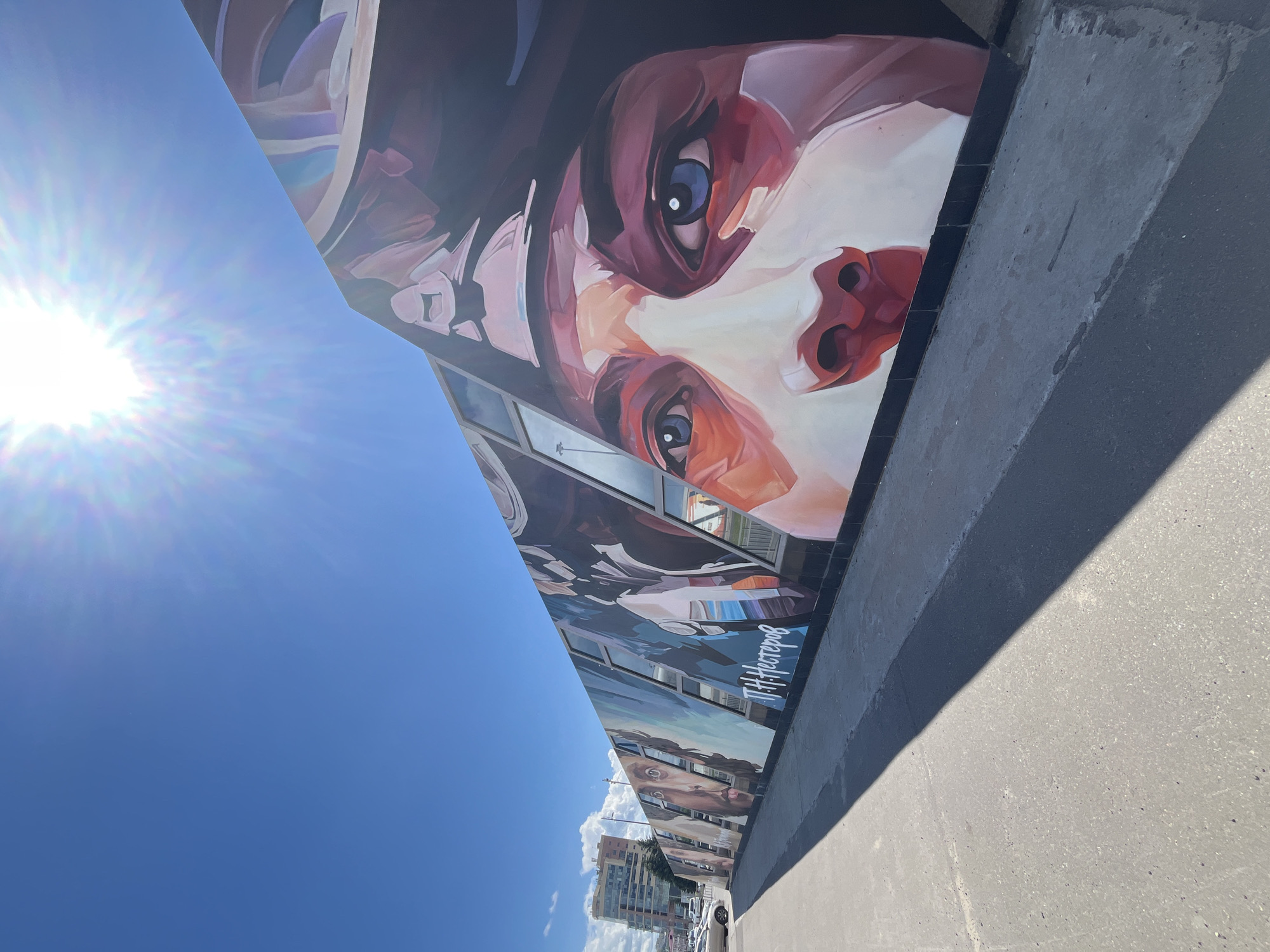 This is a wide-angle, low-angle, outdoor shot of a mural on the side of a building, with a bright sun and clear blue sky dominating the left side of the frame. The mural depicts a stylized portrait of a person, primarily focusing on their eyes and mouth, rendered in a palette of reds, browns, and whites with touches of blue in the irises. The artwork appears to be on a triangular section of the building, extending diagonally across the image. Below the main portrait, other elements of the mural are visible, including what appears to be a cityscape with buildings and cars, and more faces. Sunlight flares are present, indicating a sunny day. The lower right of the image shows a grey concrete surface, possibly a sidewalk or road, casting a distinct shadow from the mural. In the background, several apartment buildings are visible under a blue sky with scattered white clouds, suggesting an urban environment. The mural has some text written on it in white, Cyrillic script, which reads "П. Нестеров" (P. Nesterov). The overall impression is a bright, sunny day in an urban setting, highlighting a striking piece of street art.