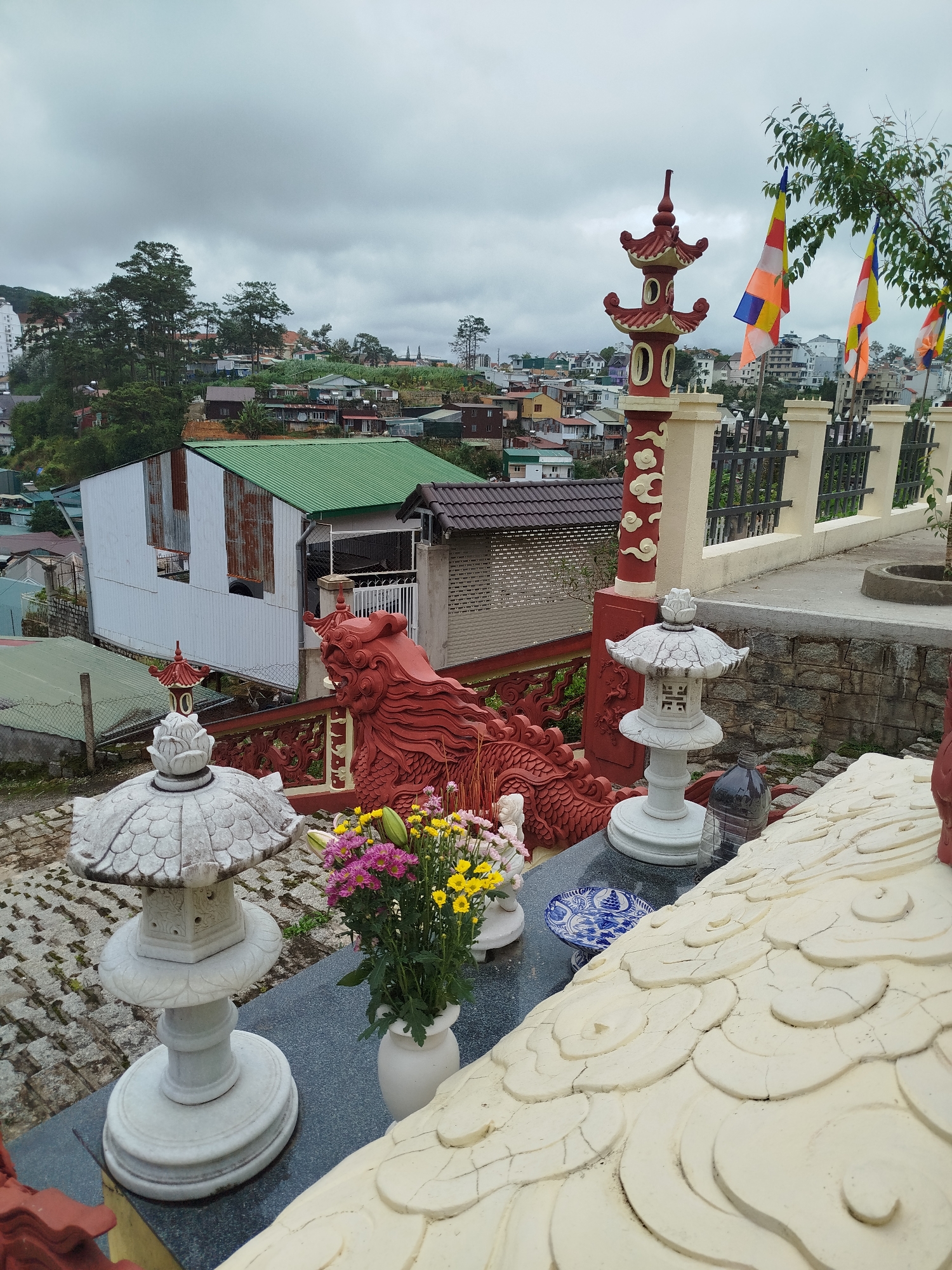 The image captures a view of a hillside urban environment in Da Lat, Vietnam, under an overcast sky. In the foreground, there are stone lantern-like structures with intricate carvings, a white vase filled with a bouquet of yellow and pink chrysanthemums, and a blue and white patterned plate. A large, red dragon sculpture is prominently featured, seemingly part of a decorative railing or wall. Behind these elements, a collection of buildings with various roof colors, including green and brown, are visible, nestled into the sloping terrain. Trees, some tall and slender, are interspersed among the structures. To the right, a beige wall with a dark metal fence separates the foreground from a pathway or elevated area. Several prayer flags, in red, yellow, blue, and orange, are strung from a pole and tree branch, adding a cultural element to the scene. The overall atmosphere suggests a temple or religious site overlooking the city.