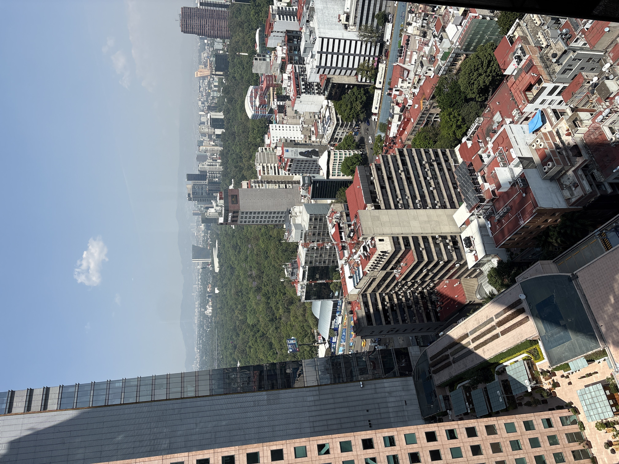 This high-angle, wide shot captures a sprawling urban landscape, likely Mexico City, stretching into the distance under a clear blue sky. The city appears vibrant and densely populated, characterized by a mix of architectural styles.
In the background, a hazy horizon reveals distant mountains, suggesting a vast metropolitan area surrounded by natural topography. The sky is bright blue with a few wispy clouds, indicating clear weather.
The mid-ground is dominated by a substantial, dense green forest or park, acting as a prominent natural divider within the urban expanse. This green space is flanked by numerous buildings, ranging from high-rise structures with modern glass facades to more traditional residential buildings often featuring red-tiled roofs. Many buildings display HVAC units and satellite dishes on their rooftops, and some modern buildings feature solar panels. A long, white truck with "Transportes Lyon & Cía." visible on its side is seen on a street running parallel to the park.
In the immediate foreground, parts of several modern buildings are visible. A large structure with dark gray cladding and expansive glass windows creates a strong diagonal shadow across its surface. Below it, a building with a reddish-pink facade and square windows is prominent. This building features several outdoor terraces and what appears to be a landscaped garden or pool area with glass enclosures, suggesting communal or private recreational spaces.
The scene depicts a sunny day, with distinct shadows cast by the buildings, indicating mid-morning to late afternoon. There are no people clearly visible due to the elevated perspective, but the bustling nature of the city is evident from the dense development and traffic. The vast green expanse amidst the urban density strongly suggests the iconic Chapultepec Park in Mexico City.