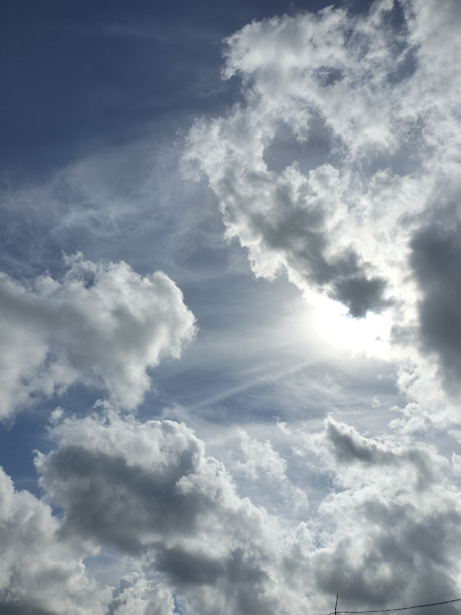 The image depicts a cloudy sky, characteristic of the climate in Karang Indah, Indonesia. The sky is filled with a mix of dark and light clouds, with patches of blue sky visible through the overcast. The sun is partially obscured by the clouds, creating a bright, diffused light that illuminates the scene. There are no people, subjects, or objects clearly identifiable in the image, indicating it is a landscape photograph focused on the natural environment. The scene is serene and natural, with the clouds creating a dynamic and textured composition.