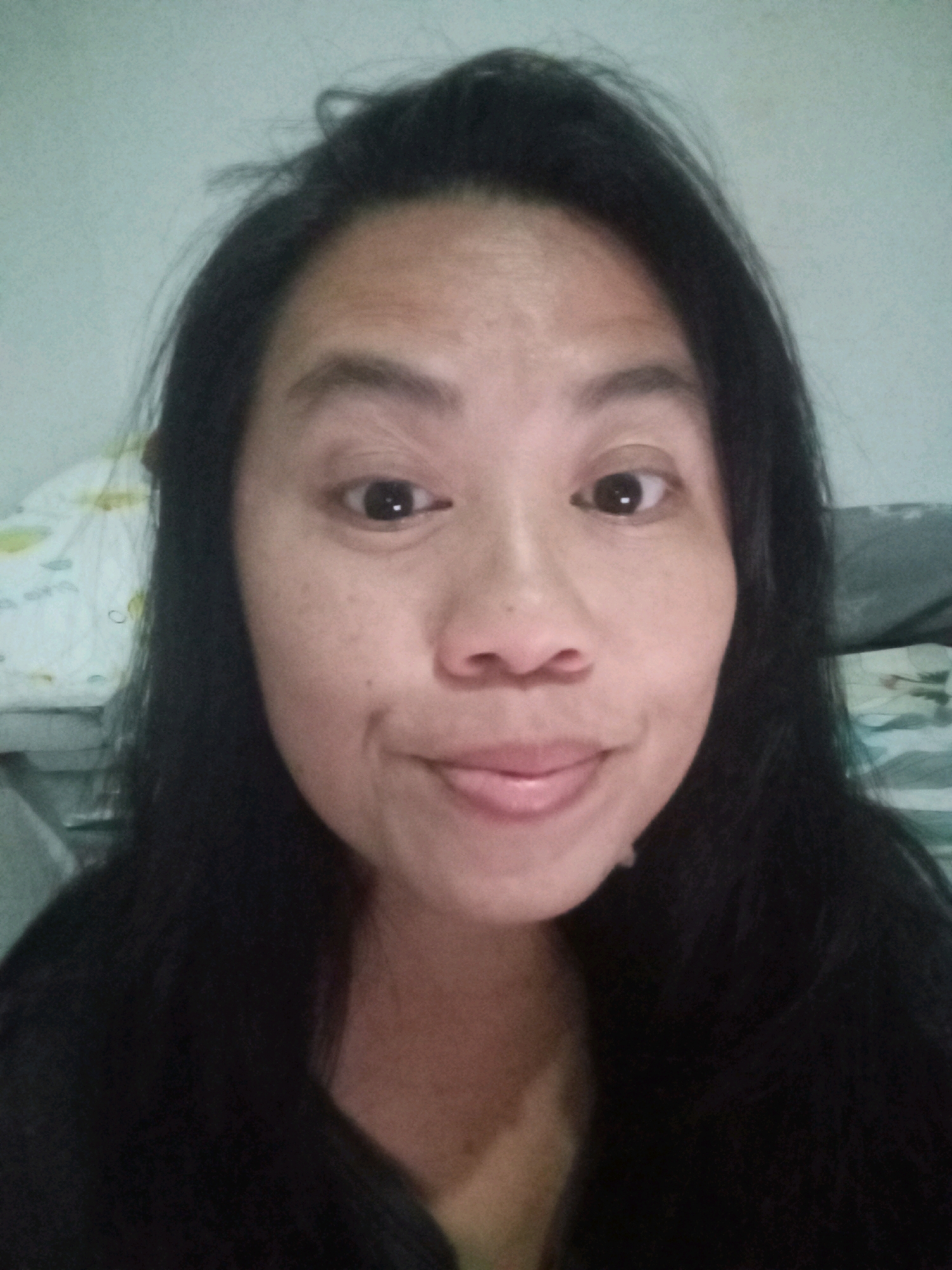 A frontal, close-up selfie of a woman with long, dark hair is captured against a pale, textured background. The woman is looking directly at the camera with a neutral expression, her mouth slightly open, and her tongue visible. Her skin is light-toned with some visible pores and slight discoloration on her forehead and cheeks. She has dark, arched eyebrows and dark eyes that appear to be looking slightly off-center. Her hair is dark and falls loosely around her face and shoulders. The background appears to be indoors, with some blurred objects visible behind her, including what looks like a patterned sheet or fabric on the left and a darker, textured surface on the right. The lighting is soft and diffused, suggesting an indoor setting with ambient light. There are no explicit indicators within the image to suggest a specific location in Denpasar, Indonesia, or any other city.