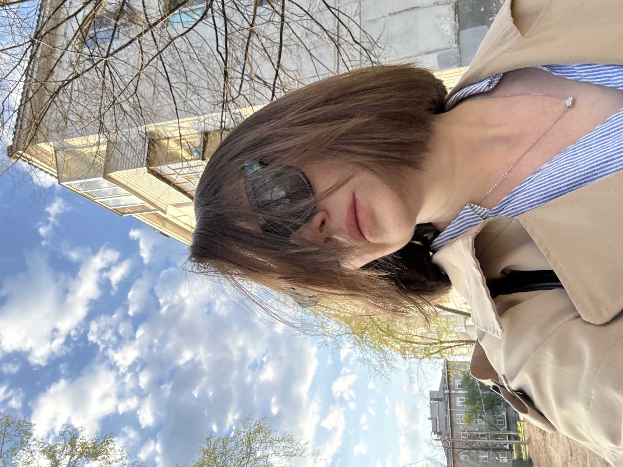 A low-angle, eye-level shot shows a person with brown hair wearing sunglasses and a beige trench coat over a blue-and-white striped shirt. A silver necklace with a heart pendant is visible. The background includes a building with beige brickwork and glass-enclosed balconies, bare tree branches, and a blue sky with scattered white clouds. Additionally, a portion of a multi-story residential building is visible. This photo was taken in the city of Chaikovsky, Russia.