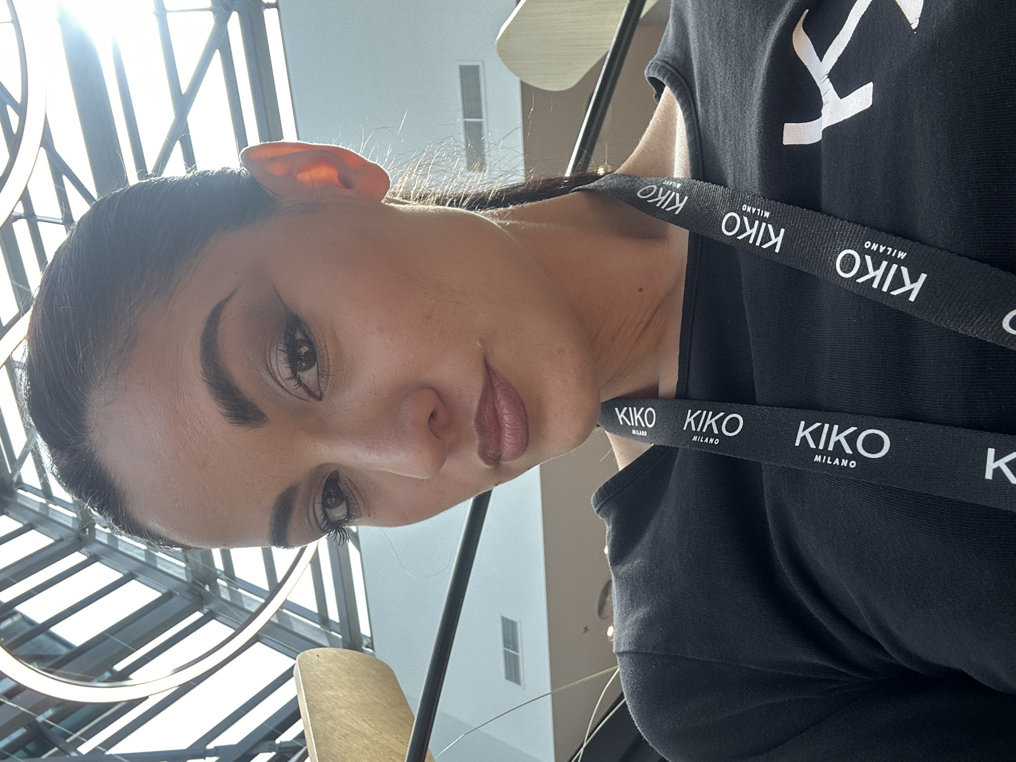 A close-up portrait shows a woman from the chest up, looking directly towards the viewer with a calm expression. She has dark hair pulled back, distinct eyebrows, and dark eyes with mascaraed lashes. Her lips are painted with a muted pink or reddish-brown lipstick. She is wearing a black top or t-shirt. Around her neck, a black lanyard is visible, featuring the brand name "KIKO MILANO" printed repeatedly in white capital letters.
The background is brightly lit, suggesting an indoor setting with abundant natural light, possibly from a large skylight or glass roof. Numerous crisscrossing beams are visible overhead, creating a grid-like pattern against the bright light. A partial view of a circular light fixture is present in the upper left, and a light-colored, possibly wooden, object is visible in the lower left background. The strong overhead lighting creates highlights on the woman's hair and skin.