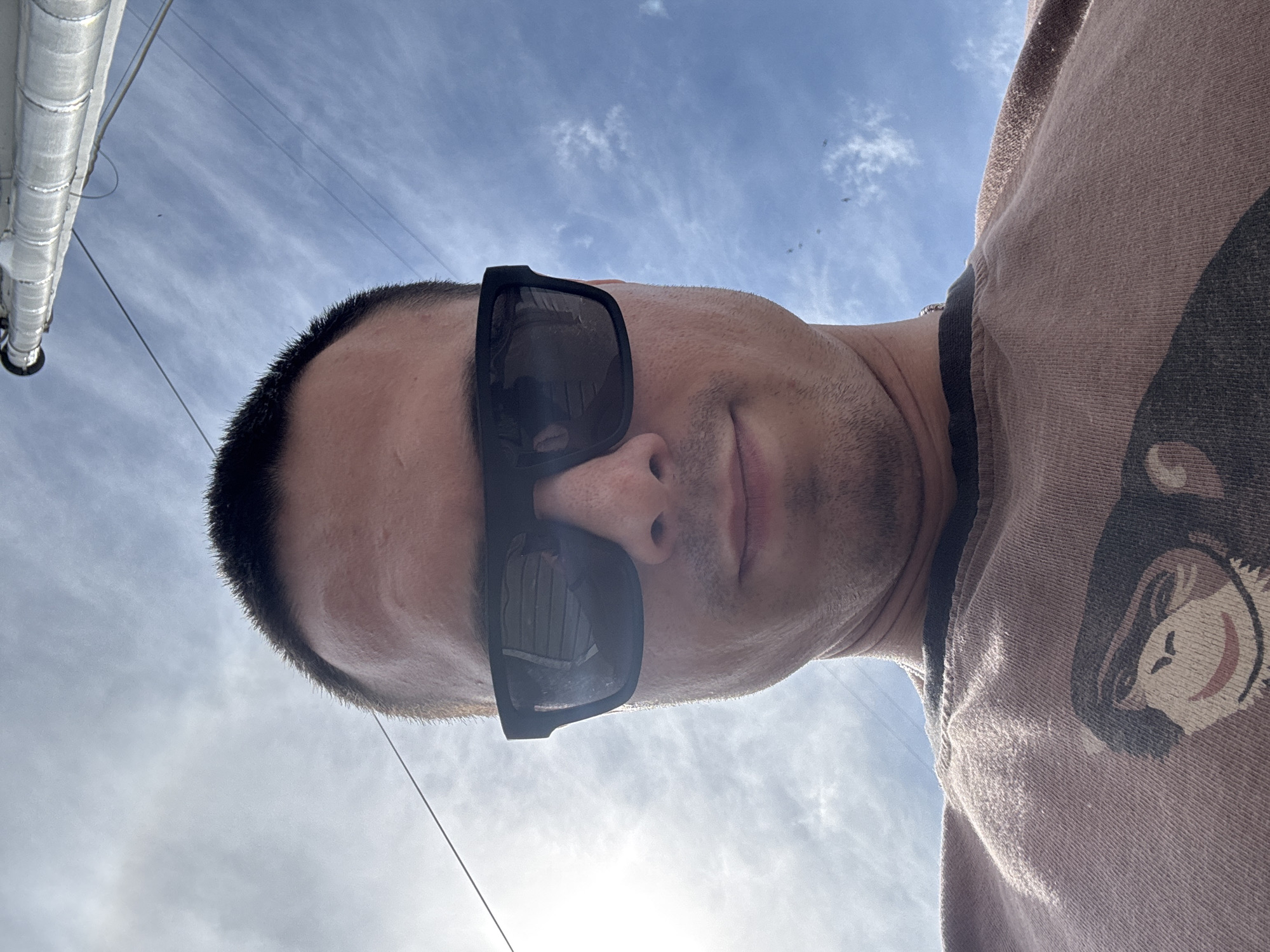 An upward-angled self-portrait shows a man wearing dark, rectangular sunglasses. The image is oriented so the man's head is on the left, looking right, with his body extending towards the right edge of the frame. He has short, dark hair and light skin, with a faint stubble visible on his chin and jawline. His lips are slightly parted.
He is wearing a light brown or tan short-sleeved t-shirt. On the right side of the shirt, a graphic of a smiling cartoon monkey's face is clearly visible.
The background is a bright blue sky with scattered, wispy white clouds. A prominent sun glare is present in the lower sky, indicating a sunny daytime. Thin, dark utility wires crisscross the upper sky. On the far left, a large, light-colored, corrugated pipe or conduit extends vertically, partially visible. The unusual perspective suggests the photo was taken from a low angle, pointing upwards, or the camera was held in an unconventional orientation. There is no visible text. The scene takes place outdoors in Chaikovsky, Russia.