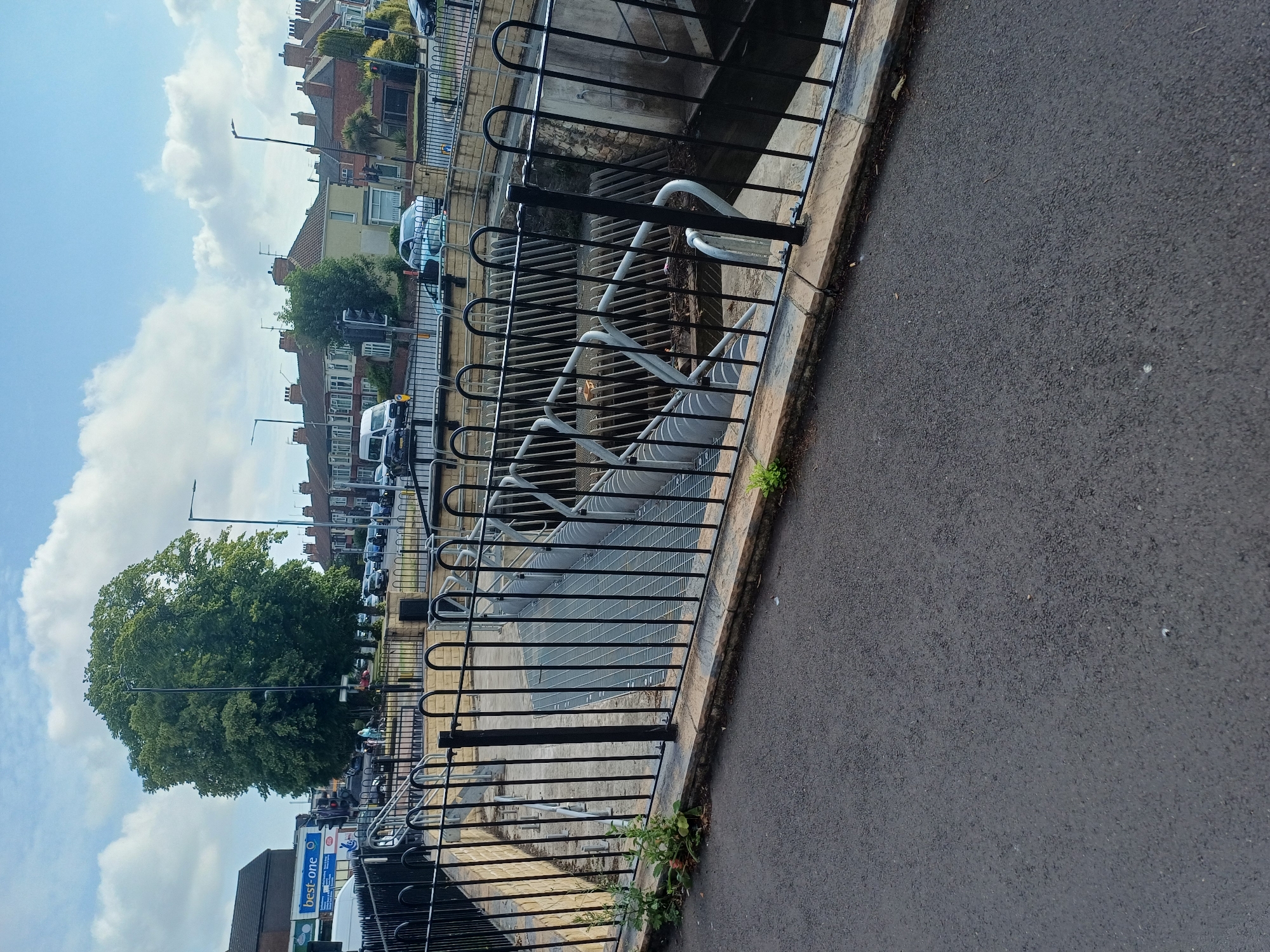 This image captures a view of the River Tees outfall structure, located near the A66 in Middlesbrough, United Kingdom. In the foreground, a paved walkway borders a black metal fence that encloses the outfall. The outfall itself is characterized by a grated metal walkway leading down into a channel. This channel contains a series of curved metal baffles, designed to regulate water flow. The background reveals a street scene with parked cars and residential buildings typical of the area. A large, leafy tree stands prominently, adding a natural element to the urban environment. The sky above is partly cloudy, suggesting fair weather.