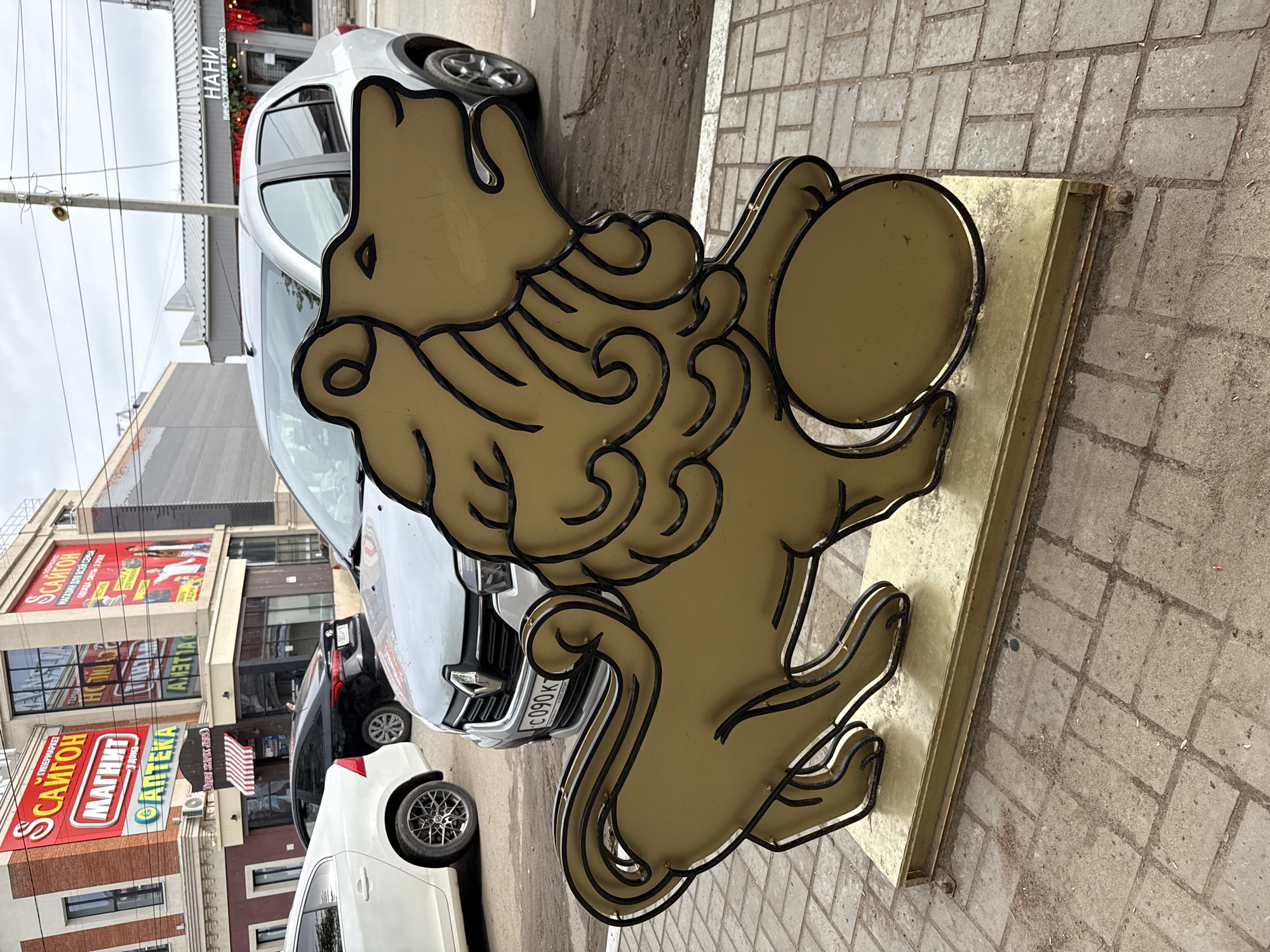 The image captures a sunny outdoor scene in Votkinsk, Russia, featuring a golden lion statue with black outlining, prominently positioned on a brick-paved surface. The lion holds a round object, possibly a shield, in its paws. Behind the statue, there are several cars, including a white Renault with license plate "C090 K" and a black car.
In the background, there is a building with multiple signs in Russian, including "САИГОН" (Saigon), "МАГНИТ" (Magnet), "АПТЕКА" (Pharmacy), and "НАНИ" (Nani), indicating a commercial area with shops and businesses. The signs suggest that the location is a busy street. A cloudy sky is visible above the buildings. The statue appears to be placed on the sidewalk, likely for promotional purposes, near the businesses.