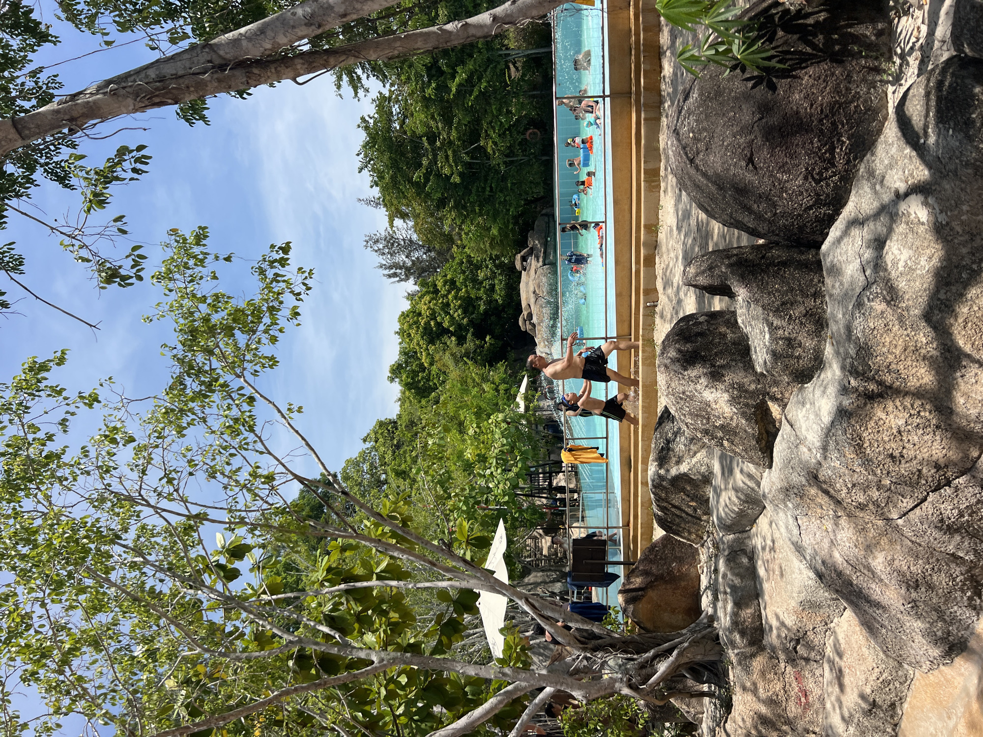 The image depicts a sunny day at a water park in Tay Nha Trang Ward, Vietnam. In the foreground, large, weathered boulders are scattered across sandy ground, casting shadows from the lush green trees and branches that frame the scene. Beyond the boulders, a bright blue swimming pool is visible, surrounded by an orange-tiled deck. Several people are enjoying the water, including children and adults, with some floating on inflatable rings and others swimming. Two men are walking near the edge of the pool, one in black shorts and the other in patterned swim trunks. In the background, dense green trees create a natural backdrop, with a clear blue sky overhead. White umbrellas are set up near the pool, suggesting areas for shade and relaxation.