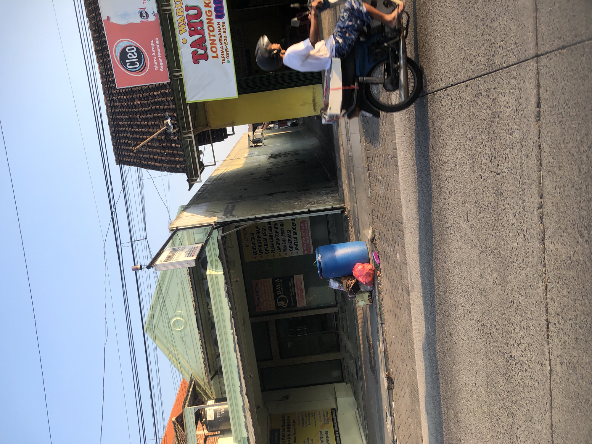 This is a daytime, eye-level, medium shot of a street scene in Boro, Indonesia. The foreground shows a gray asphalt road with a visible seam running through it, and a shadow cast from the right. In the midground, a person wearing a white t-shirt and patterned shorts rides a blue motorcycle, viewed from the right side. The motorcycle has a white box on the back. The person is wearing a black helmet and appears to be looking forward. To the left of the motorcycle, a paved sidewalk is visible with several trash bags and a large blue plastic barrel. Behind the sidewalk, there is a building with a light green facade, an open doorway, and several signs with text in Indonesian. One sign advertises "WARUNG TAHU LONTONG KI". Further to the right, another building has a brown tiled roof and a large red and white billboard advertising "Cleo" bottled water. The sky is clear and light blue. The overall impression is a busy street with commercial establishments and the typical elements of a residential or commercial area in Indonesia.