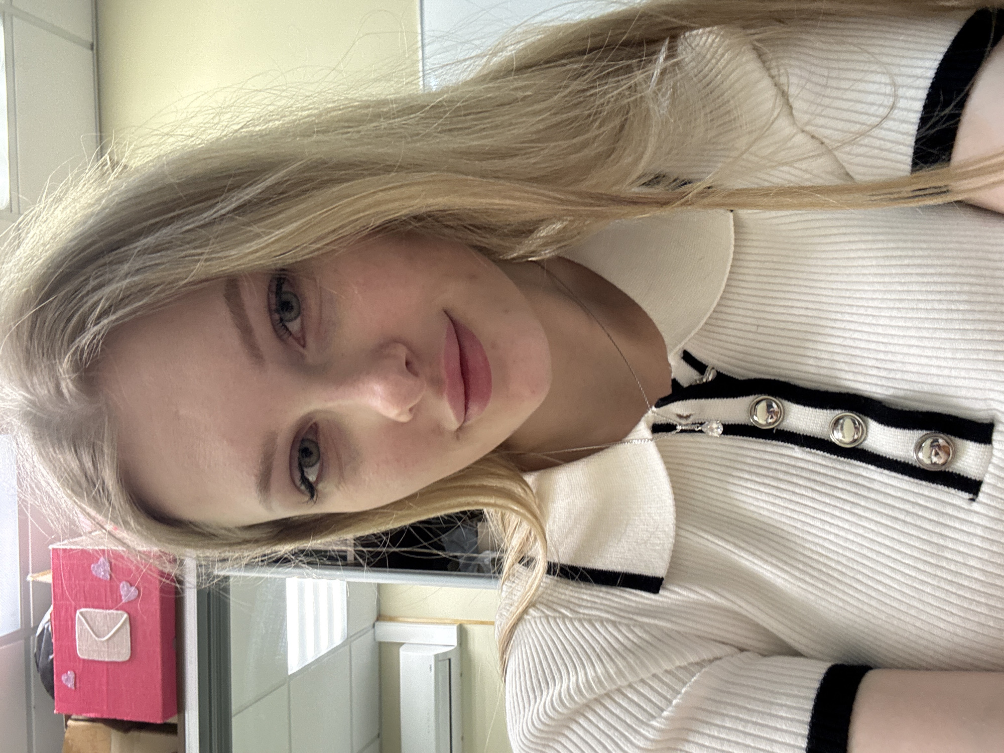 A blonde woman poses indoors. She wears a white collared shirt with black trim and silver buttons. A silver necklace adorns her neck. A pink box with an envelope design and heart decorations sits on a shelf behind her, near a tiled wall and window. The location is Perm, Russia.
