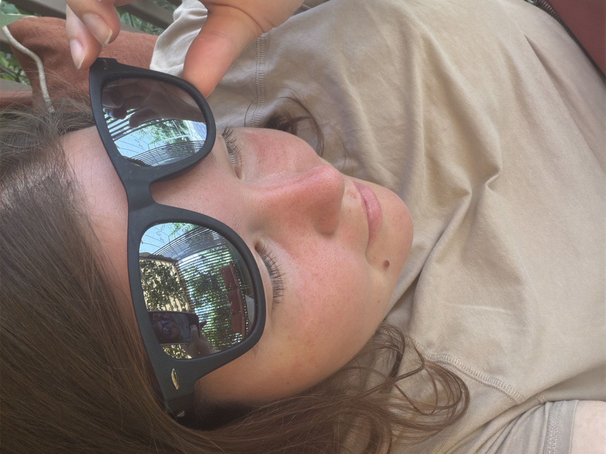 The image is a close-up, high-angle shot of a person's face and upper torso, taken outdoors. The subject, wearing sunglasses, is looking down and to the right. The sunglasses reflect a scene of outdoor seating with red chairs and a fence, suggesting a public or semi-public outdoor space. The person's skin shows some redness, possibly from sun exposure, and a mole is visible on their cheek. The person is wearing a beige t-shirt. The background is blurred, with hints of greenery and what appears to be a light-colored awning or roof structure. The image does not contain any discernible details that would suggest a specific location within Bishkek City, Kyrgyzstan.