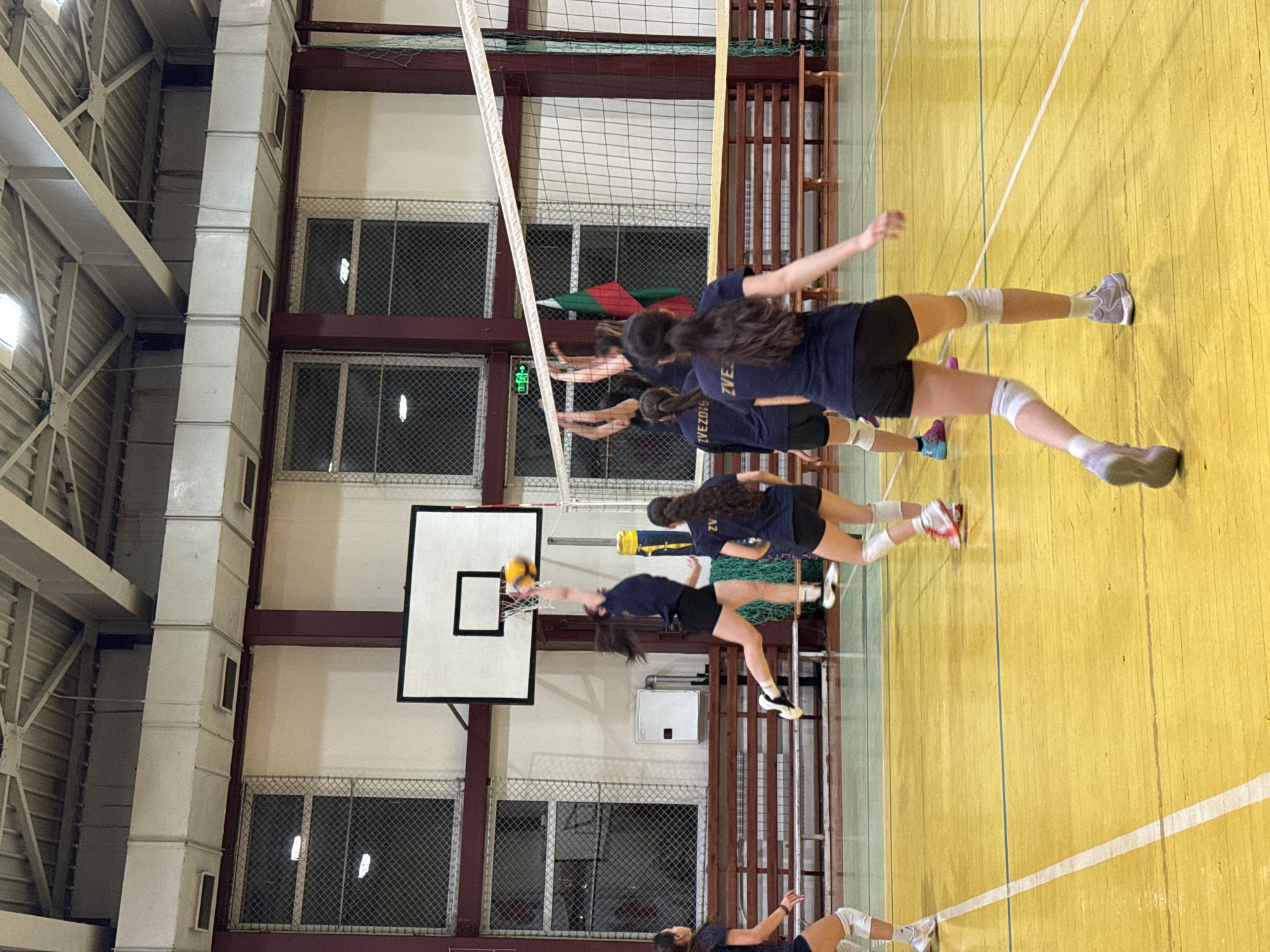 The image shows a volleyball game in progress inside a gymnasium. Several female volleyball players are positioned near the net. They are wearing dark blue jerseys with the word "ZVEZDA" printed on them, along with black shorts and knee pads. One of the players is spiking the ball. The gymnasium has a yellow wooden floor with white boundary lines, and a basketball hoop is visible on the wall. The gymnasium has a network of maroon and beige-painted metal beams. The ceiling has exposed ductwork. Tall windows with mesh coverings are built into the walls. The presence of the "Zvezda" team suggests the location is likely in Sofia, Bulgaria, as "Zvezda" means "star" in Bulgarian and is a common name for sports teams in the area.