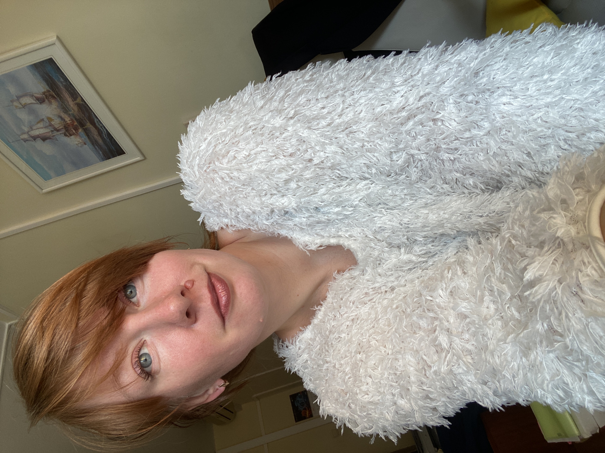 A close-up, rotated portrait captures a young woman with shoulder-length red/auburn hair and light blue eyes. She looks directly at the camera with a soft, gentle smile, her lips glossed. She is wearing a distinctly fluffy, shaggy white garment, possibly a sweater or jacket, which has a textured, almost feather-like appearance. A small gold earring is visible on her right ear.
The setting is indoors, with a light-colored wall serving as the background. On the left side of the frame, a framed picture is visible, depicting a large sailing ship on a blue sea under a partly cloudy sky. To the right and behind the woman, indistinct dark furniture, possibly the backrest of a chair or sofa, and a hint of a yellow object are present. The lighting is soft and natural, suggesting it is daytime. The scene is located in Krasnoyarsk, Russia. No visible text is present.