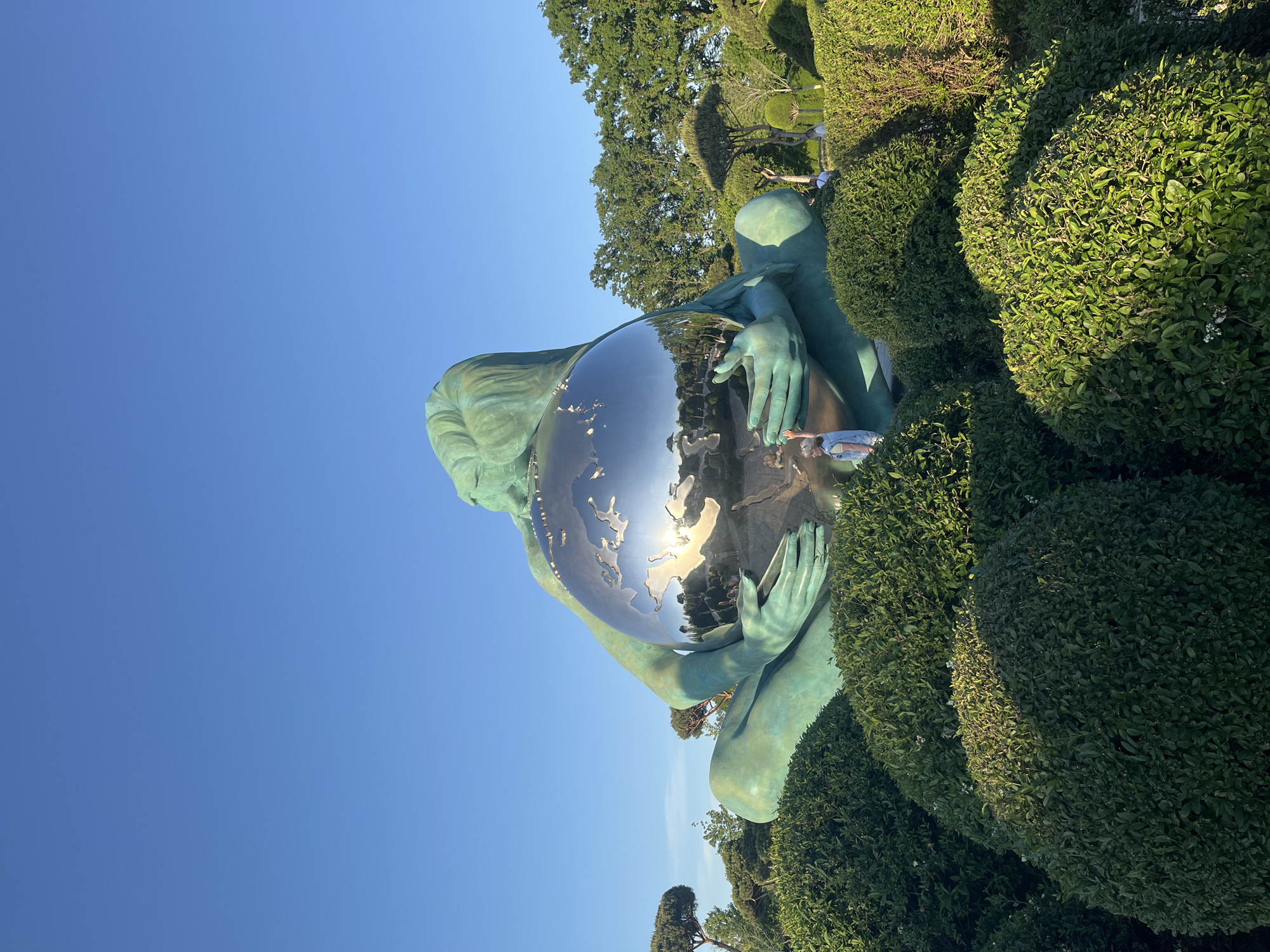 A large, verdigris-patinated sculpture of a figure, possibly a woman, gracefully holding a spherical globe, is depicted in an outdoor setting under a clear blue sky. The image is rotated counter-clockwise. The sculpture dominates the mid-ground, featuring intricate details on the figure's hands and flowing hair.
The globe held by the figure is highly reflective and polished, acting as a mirror that captures the surrounding environment. Prominently visible on its surface are the continents of Europe and Africa, along with a bright, intense reflection of the sun, indicating clear and sunny daytime weather.
The sculpture is surrounded by lush, dark green, manicured hedges and larger trees, suggesting a well-maintained park or garden. A small person, possibly a child, is visible at the base of the sculpture on the right, appearing to touch or interact with it. The scene conveys a sense of artistic integration with nature, characteristic of a public garden or specialized park in Krasnodar, Russia. No discernible text is visible in the image.