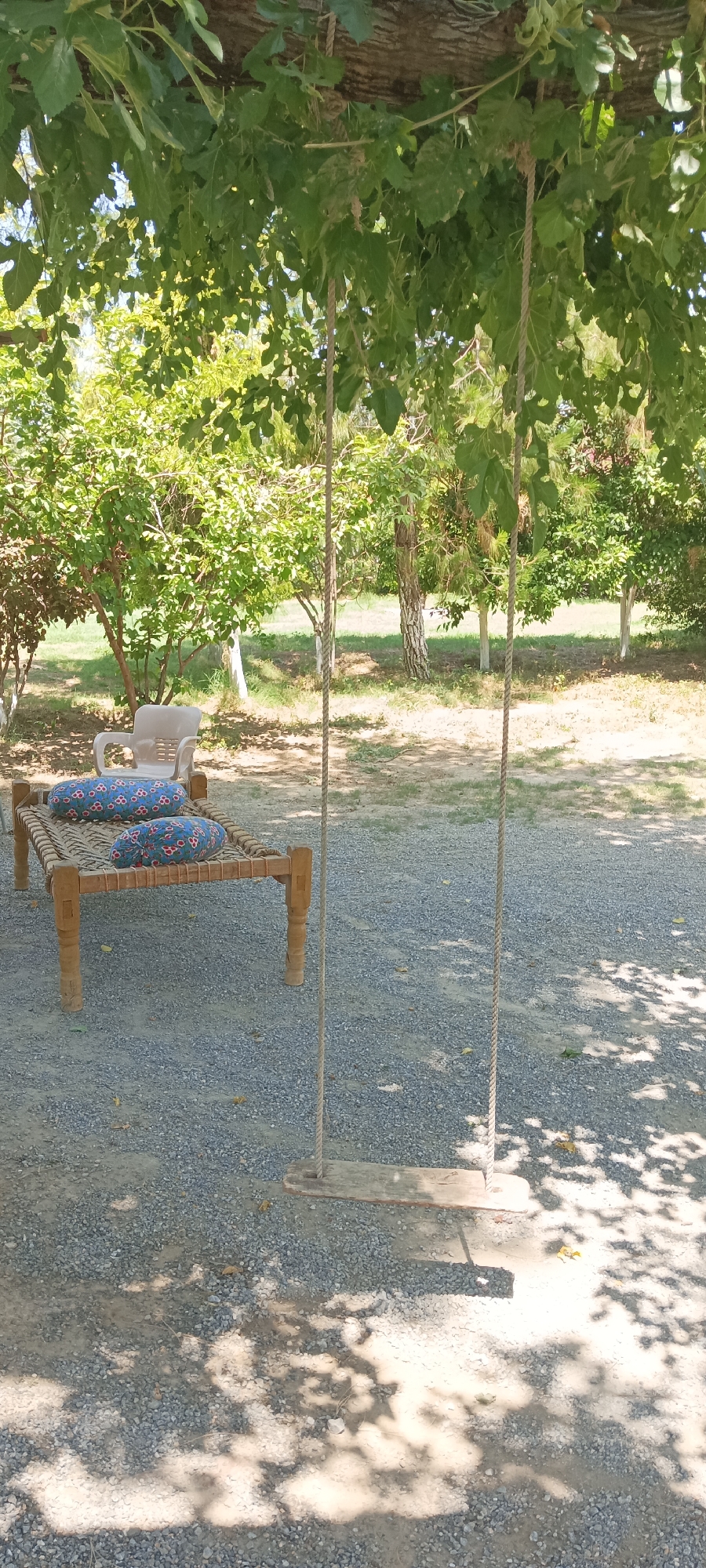 The image depicts a shaded outdoor scene in Alam Khan Kalay, Pakistan. A traditional wooden bed with two patterned, cylindrical pillows rests on a gravel surface. A white plastic chair sits nearby. A swing hangs from a tree branch above, casting shadows on the ground. Various trees and foliage fill the background.