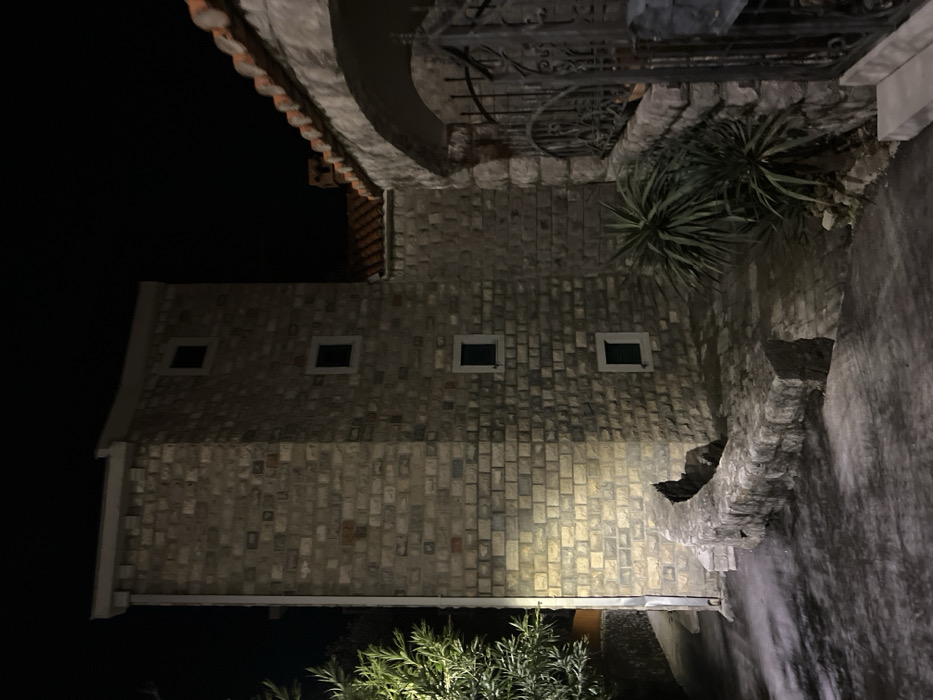The image displays a stone-clad building at night in Budva, Montenegro. The central structure is a multi-story tower-like edifice composed of irregular light-colored stone blocks. Four small, rectangular windows with dark frames are visible on its primary face; the lower three appear to have closed dark shutters. A light-colored vertical downspout is affixed to the left corner of this tower. To the right, an adjacent stone-clad section of the building features an arched entrance leading to a dark wrought-iron gate. A terracotta-tiled roof with a visible chimney extends above this section.
The foreground consists of a dark paved surface exhibiting lighter patches where artificial light falls. A low, curved stone wall, constructed from similar stone to the building, demarcates the paved area from a slightly elevated terrain. Green, leafy vegetation is present on the far left, illuminated by a direct light source. Several spiky, succulent-type plants are visible in the elevated area near the curved stone wall and the arched entrance. The scene is illuminated by artificial light, casting distinct shadows on the ground and building surfaces. No individuals or discernible activities are present.