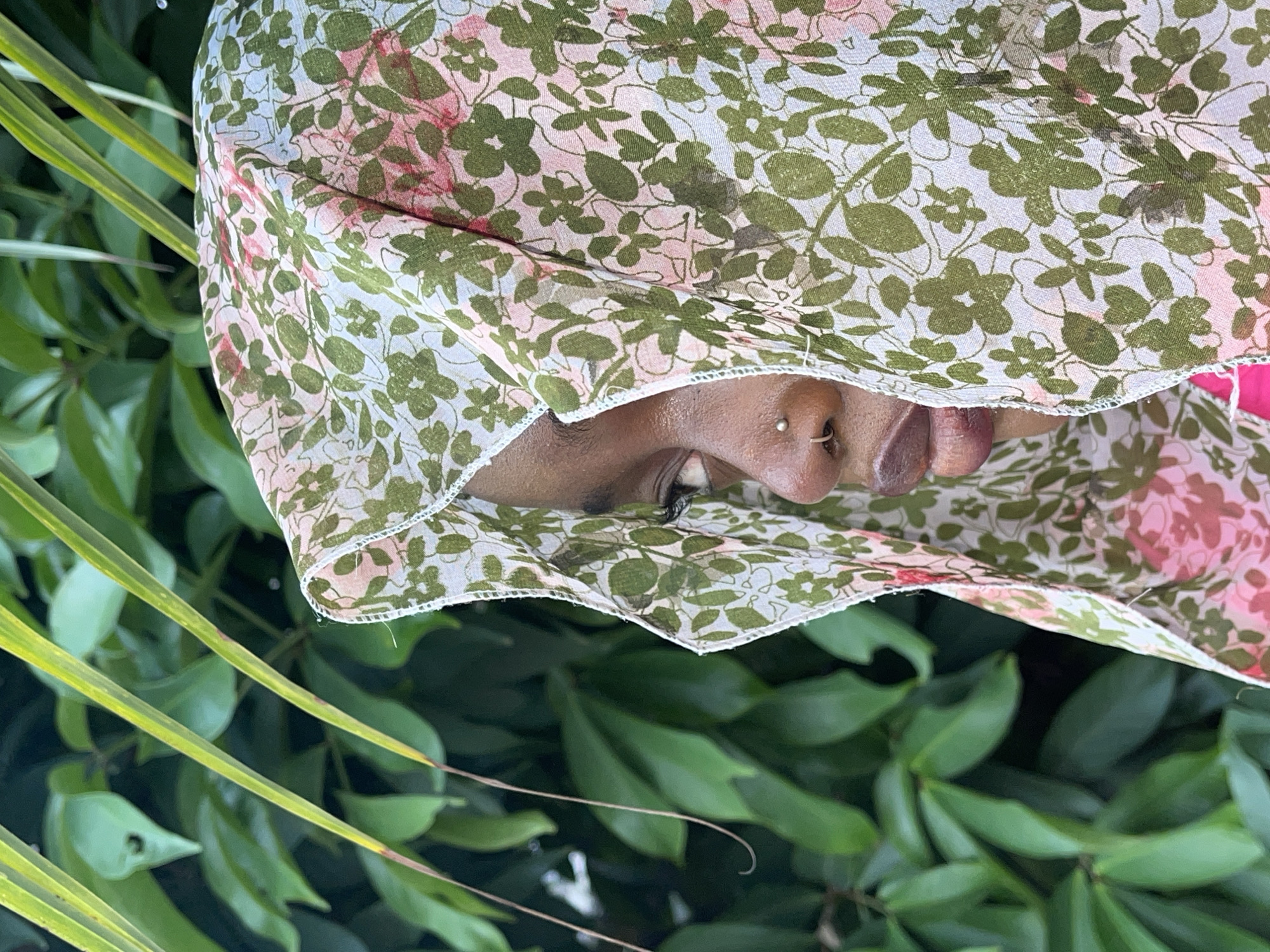 The media file is a close-up, rotated 90 degrees clockwise, depicting a person partially obscured by a patterned fabric against a backdrop of lush green foliage. The visible portion of the person's face shows dark skin, a prominent eye with visible eyeliner or mascara, a nose with a small silver stud piercing in the nostril, and part of the lips, which appear full and possibly glossed.
The person is peeking from under or looking out from behind a light, sheer fabric. This fabric features a vibrant pattern of green leaves and clusters of pink blossoms on a white or light blue background, suggesting a floral or botanical design. The edge of the fabric, with its visible white stitching, frames the lower part of the visible face.
The background is a soft-focus blur of dense green leaves and plants, indicating an outdoor setting with abundant vegetation, possibly a garden or forested area. The bright, natural lighting suggests it is daytime. No specific activity beyond posing or observing can be definitively identified. The overall impression is one of calm or contemplation, with the patterned fabric adding a decorative element. No text is visible in the image. The location context provided is Ado, Nigeria.