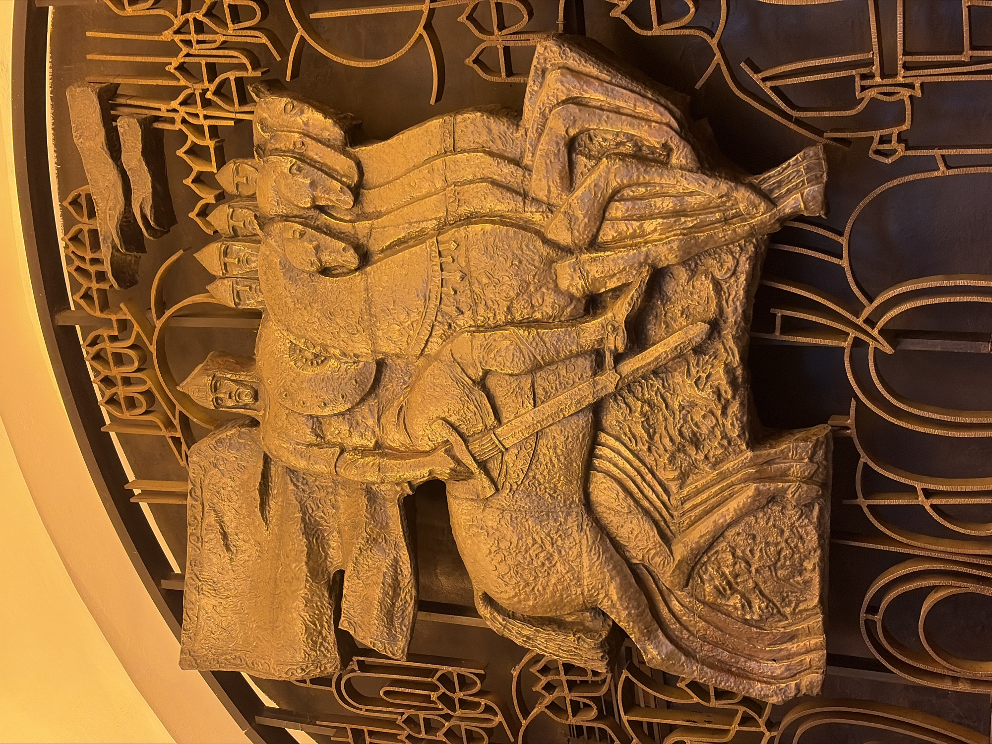 A low-angle, close-up shot depicts a gold-colored, three-dimensional bas-relief sculpture mounted on a darker-colored, patterned metal backdrop. The sculpture features an equestrian scene, where a rider, armed with what appears to be a sword or lance, is mounted on a rearing horse. The rider appears to be in the midst of battle, possibly leading a charge or engaged in combat. Several helmeted figures are positioned on the left side of the equestrian group. The entire sculpture is highly textured, with visible patterns. A wall, colored in a warm yellow hue, is barely visible on the left side of the frame. This bas-relief can be found in the Saint Petersburg Metro, Russia.