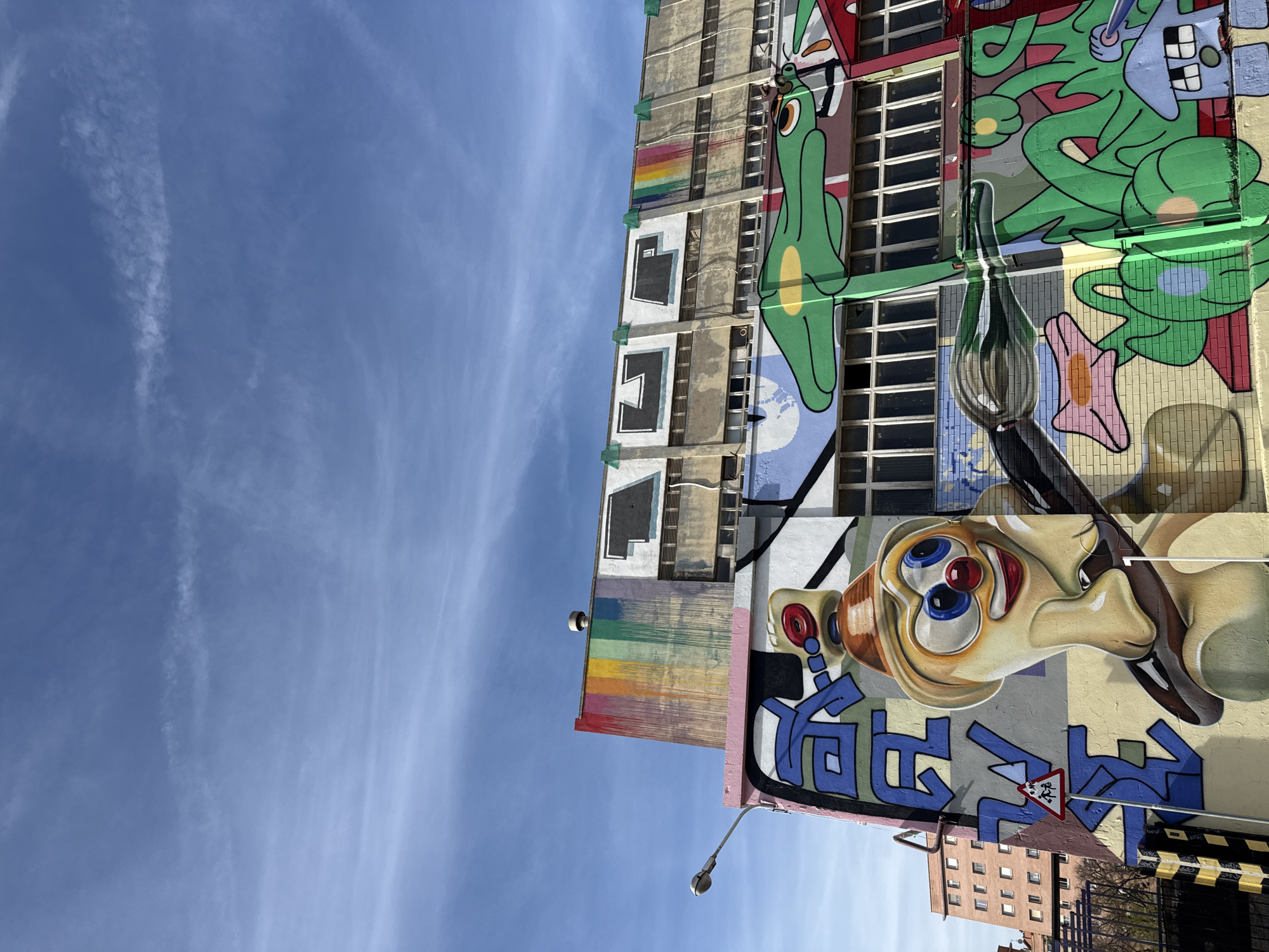 An eye-level, vertical shot shows a building in Barcelona, Spain, covered in cartoonish graffiti with a clear blue sky overhead.
The building is several stories high and appears to be made of concrete. Its exterior is painted with a variety of colorful graffiti, including cartoon characters, abstract designs, and geometric shapes. A large, elongated, dog-like face, rendered in shades of beige, brown, red and blue, is painted at the lower-right side of the shot. Above it are green cartoonish figures. A rainbow is also painted near the top-left edge.
The background consists of a clear blue sky with wispy clouds. A street light is visible at the lower-left and a building is visible near the lower-right.