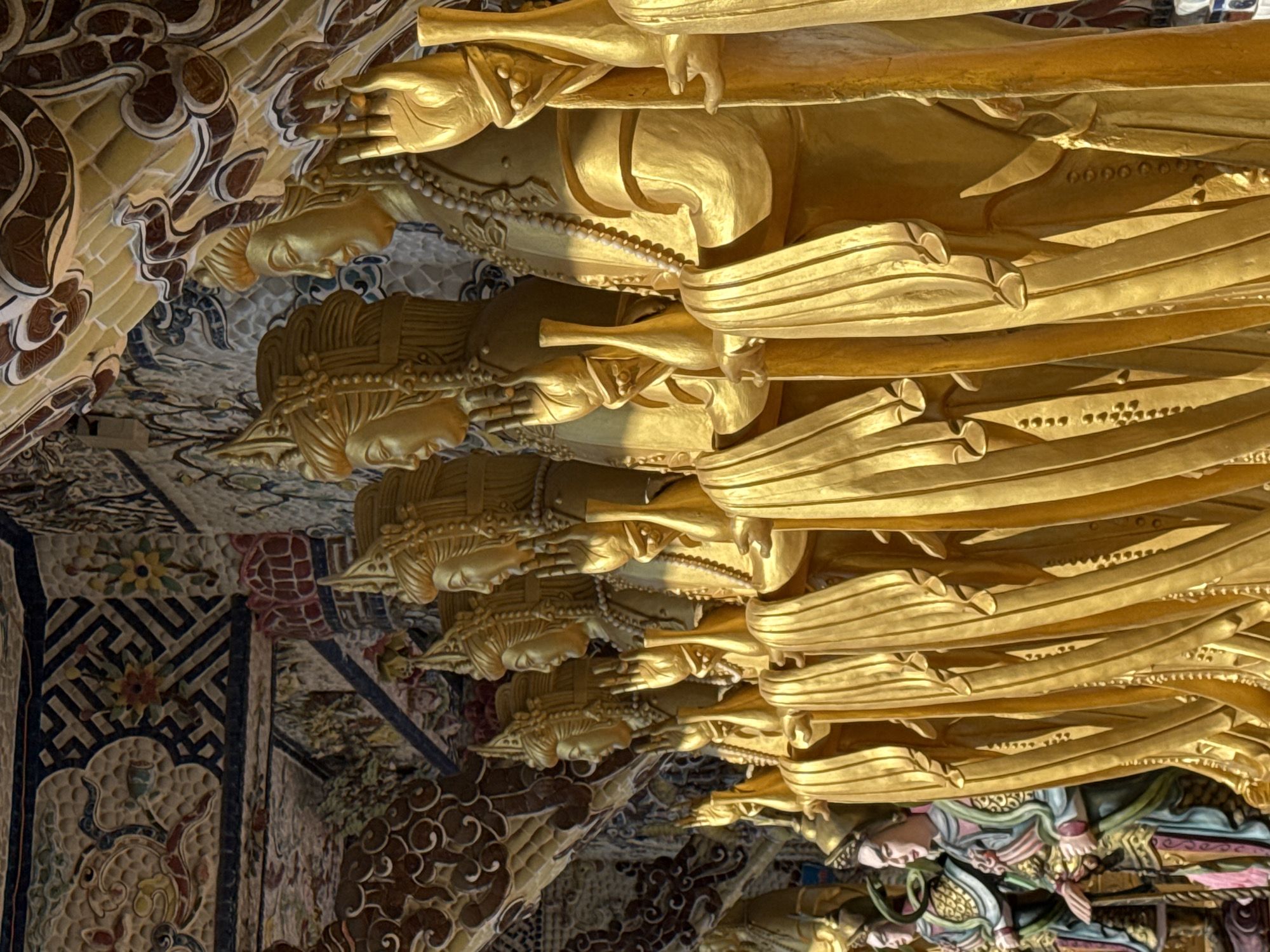 In Xuân Thành, Vietnam, a row of gilded statues stands in front of a decorative wall and ornate ceiling. The statues, depicted with serene expressions and elaborate headdresses, are adorned with beaded necklaces and draped robes. They are positioned in a line, facing left, with their hands raised in a gesture of blessing or protection. The wall behind them is covered in colorful mosaics and intricate patterns, while the ceiling features sculpted clouds and decorative motifs. The lighting suggests the scene is captured outdoors, with sunlight illuminating the golden statues.