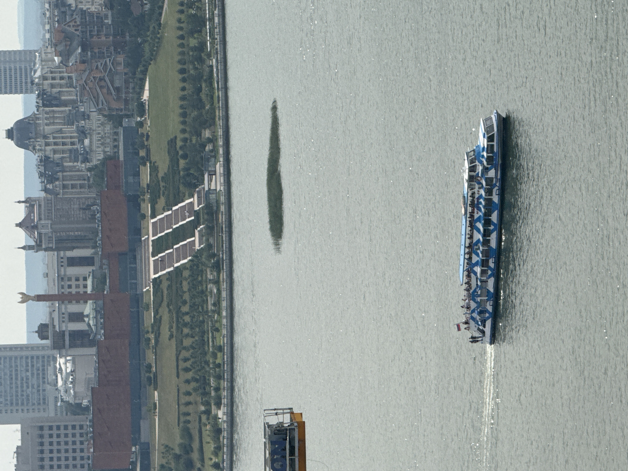 The image shows a wide view of the Kazanka River in Kazan, Russia. In the foreground, a blue and white passenger boat with numerous people on board cruises across the water, leaving a wake. To the left, a yellow structure, possibly a construction crane or a viewing platform, extends into the frame over the water. In the mid-ground, a cluster of dark green reeds forms a small island in the river. The background features the city skyline of Kazan, dominated by a large, reddish-brown building with a grand staircase leading up to it. Further in the distance, various architectural styles of buildings are visible, including tall modern structures, ornate older buildings with domes, and brick edifices. The overall scene depicts a section of the city's waterfront with a river cruise underway, framed by urban architecture and natural elements.