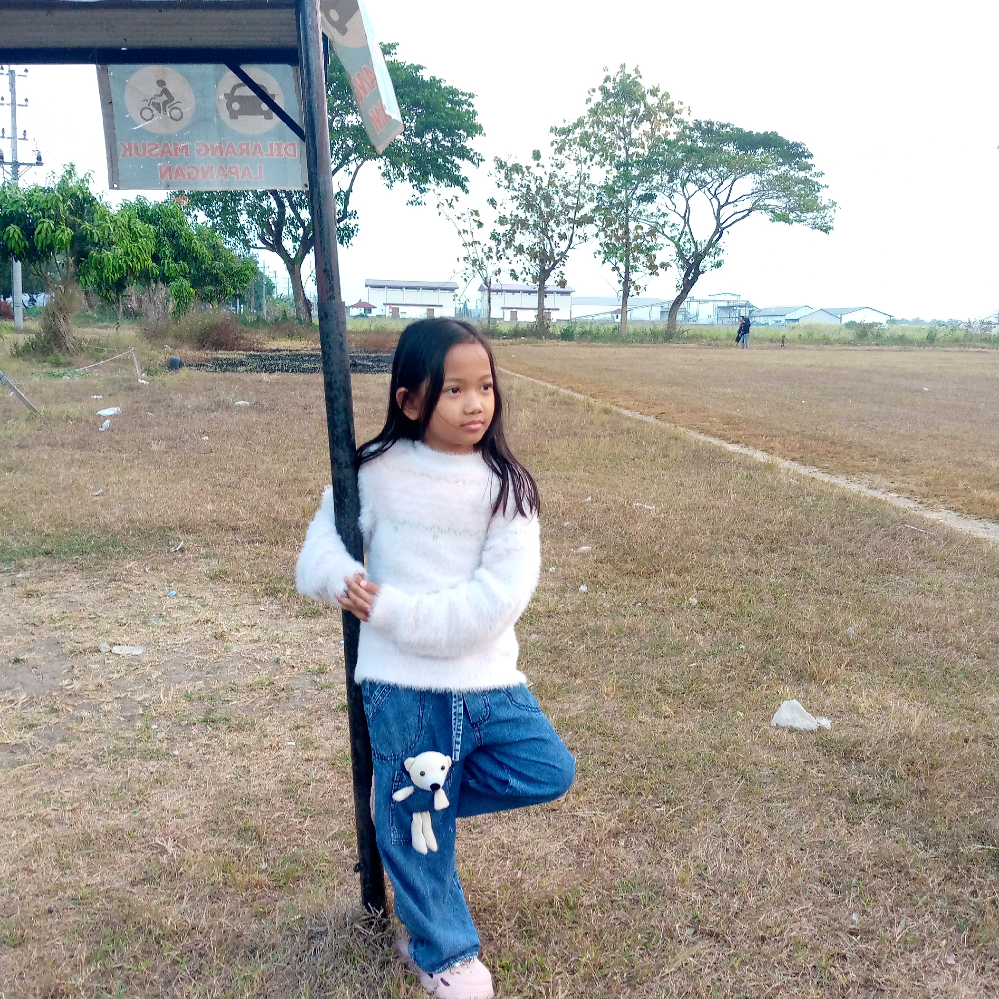 The image shows a young girl standing in a dry, grassy field, likely a sports field, in Pacing, Indonesia. She is leaning against a dark pole, wearing a fluffy white sweater and blue jeans. A small white teddy bear is attached to her jeans. In the background, there are trees, buildings, and a sign with pictograms indicating "No motorcycles" and "No cars". The overall impression is of an outdoor scene in a somewhat unkempt environment.