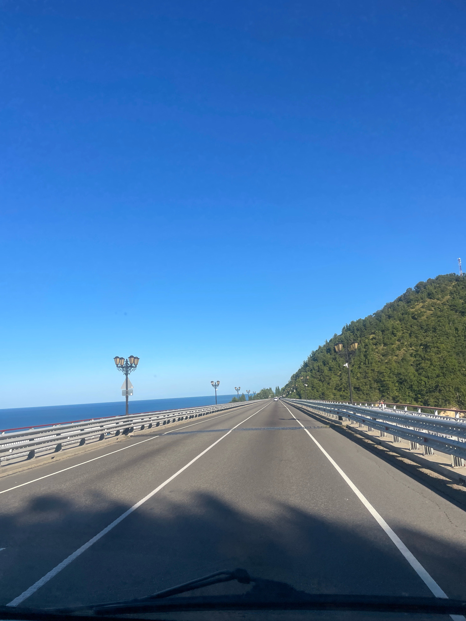 The image captures a view from a moving vehicle on a highway, likely in Sochi, Russia, given the scenic coastal road and mountainous backdrop. The highway stretches forward, bordered by a metal guardrail and an expanse of blue ocean on the left. A steep, tree-covered mountain rises on the right, with a communication tower visible at its peak. Several ornate lampposts line the roadside. The sky is a clear, vibrant blue, indicating fair weather. Shadows of trees and possibly overhead structures are cast on the asphalt road, suggesting a sunny day. No people are clearly visible in the frame, though the presence of the road and guardrail implies human infrastructure. The overall scene depicts a picturesque coastal drive.