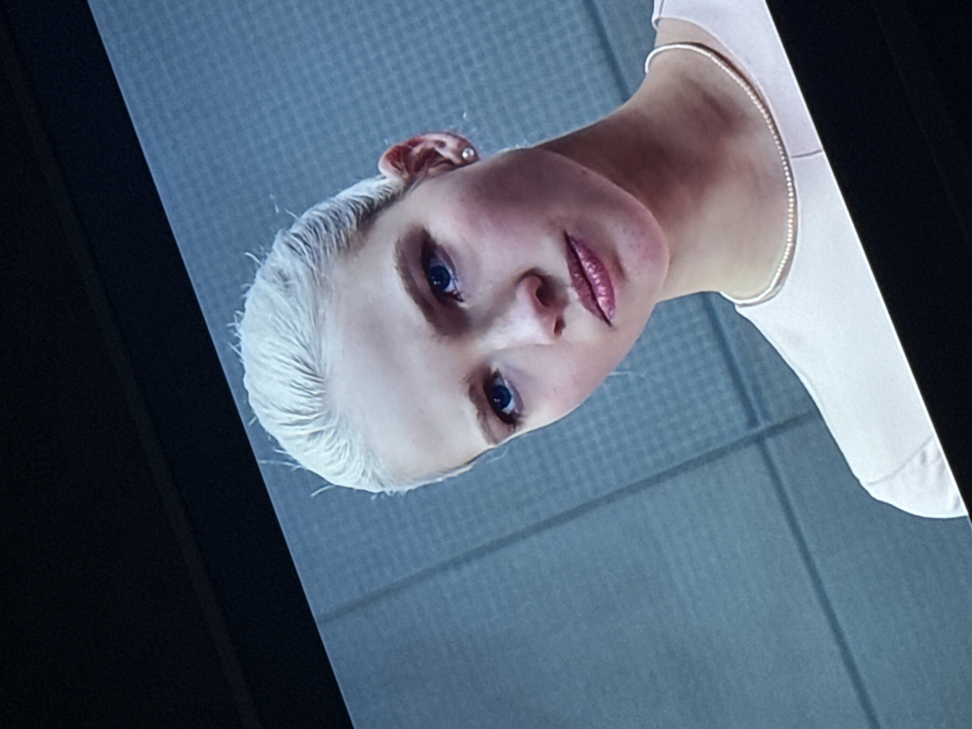 A close-up, rotated view captures a woman with short, bleached blonde hair. She has fair skin, blue eyes, and is wearing pink lipstick. She wears a delicate silver necklace and a white garment that partially covers her shoulder. The background is a textured, light blue-gray. The image is framed by what appears to be a dark border, possibly a monitor or picture frame. Location details are not discernible from this image.