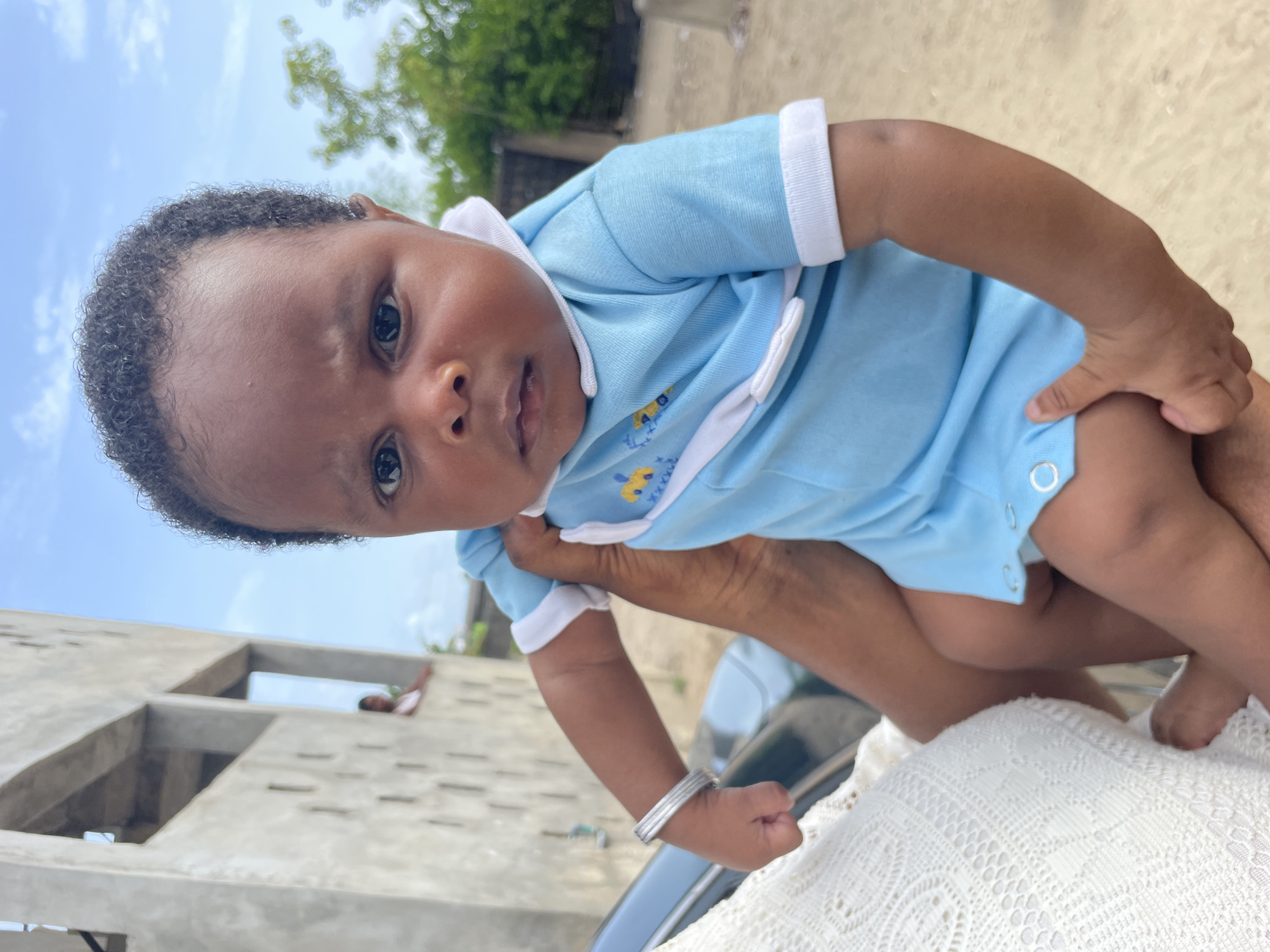 A baby wearing a light blue outfit with white trim is held in someone's arms. The baby has dark skin, dark curly hair, and dark eyes. The person holding the baby has brown skin and is wearing a silver bracelet. A white crocheted blanket is partially visible. The background includes a building under construction, vegetation, and a light blue sky with clouds. The location is Badagry, Nigeria.