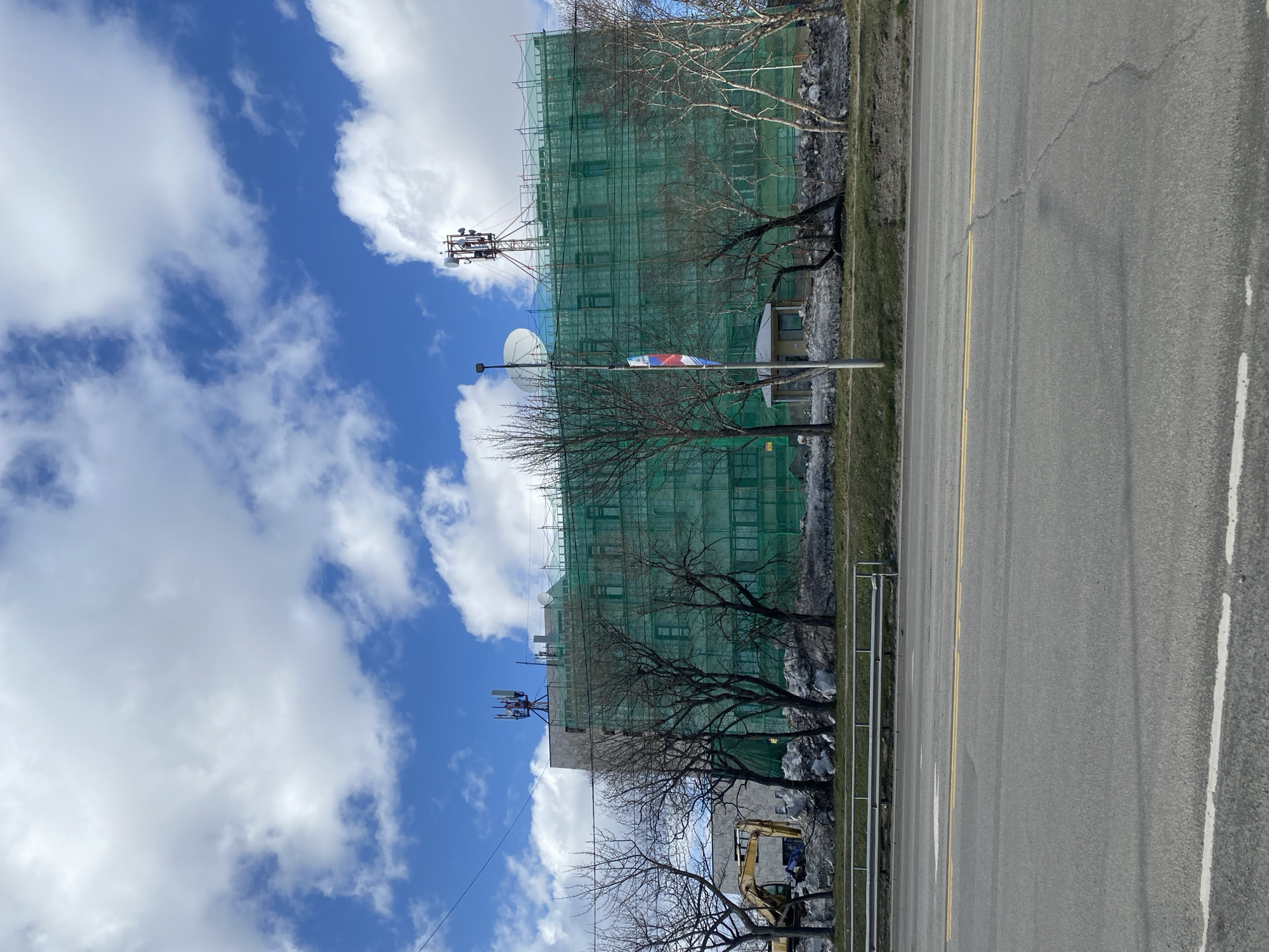 The image depicts a street scene in Petropavlovsk-Kamchatsky, Russia, adjacent to a multi-story building undergoing construction or renovation. The building is covered in green scaffolding netting. A flagpole with a flag is in front of the building, and communication towers and satellite dishes are mounted on the building's top. Bare trees are scattered around the building's base. A two-lane road with yellow center lines is visible in the foreground. The sky is bright blue with scattered white clouds. Construction equipment is parked near the road.