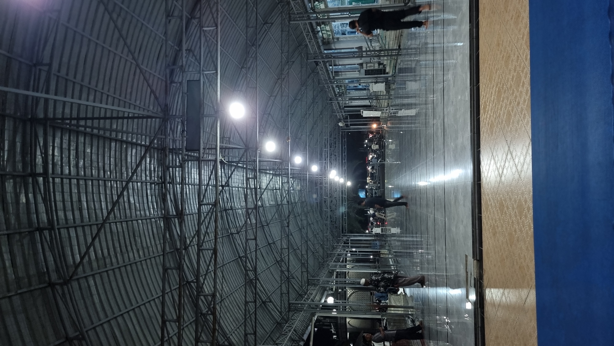 The image depicts an open-air covered structure with a metal roof supported by numerous metal beams and columns. The floor is highly polished and reflects the overhead lights and the few people present. Several individuals are visible, appearing to be walking or standing in the space. Some are dressed in traditional attire, including head coverings. The environment suggests a place of gathering or worship, possibly a mosque courtyard given the context of Keudekruenggeukueh, Indonesia, a region known for its Islamic heritage. The lighting is artificial, from overhead lamps, and casts a cool tone across the scene. The time of day appears to be evening or night due to the darkness in the background and the prominent artificial lighting. The overall atmosphere is serene and functional.