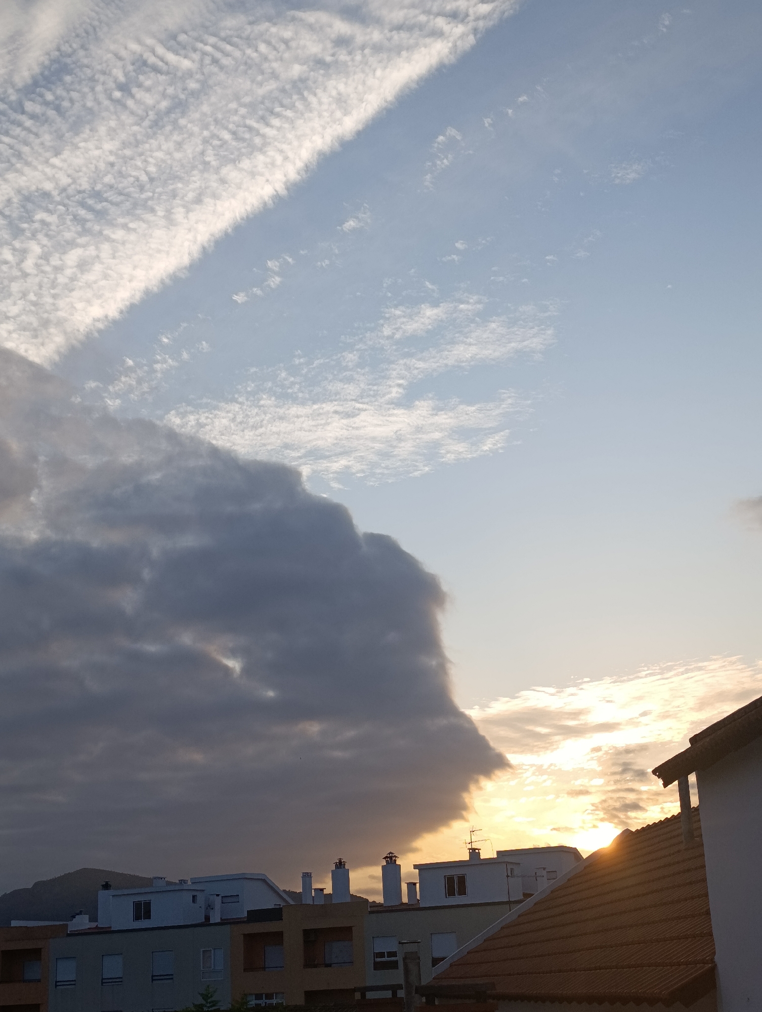 The image captures a dramatic sky over the city of Setúbal, Portugal, during what appears to be late afternoon or early evening. The sky is divided by a sharp, diagonal band of dark, dense clouds that dramatically contrasts with the lighter, scattered cirrus clouds above and the bright, sunlit sky to the right. The sun is low in the sky, casting a warm, golden glow that is partially obscured by the larger cloud formations, creating areas of intense brightness and shadow.
In the foreground, the rooftops of several buildings are visible. These appear to be residential buildings, characterized by tiled roofs, chimneys, and visible window frames. The architectural style suggests a European urban setting. The buildings are clustered together, indicating a densely populated area. A hill or mountain range is faintly visible in the background, silhouetted against the dimmer part of the sky. There are no people or vehicles clearly identifiable in the image. The overall impression is one of natural atmospheric drama occurring over an urban landscape.