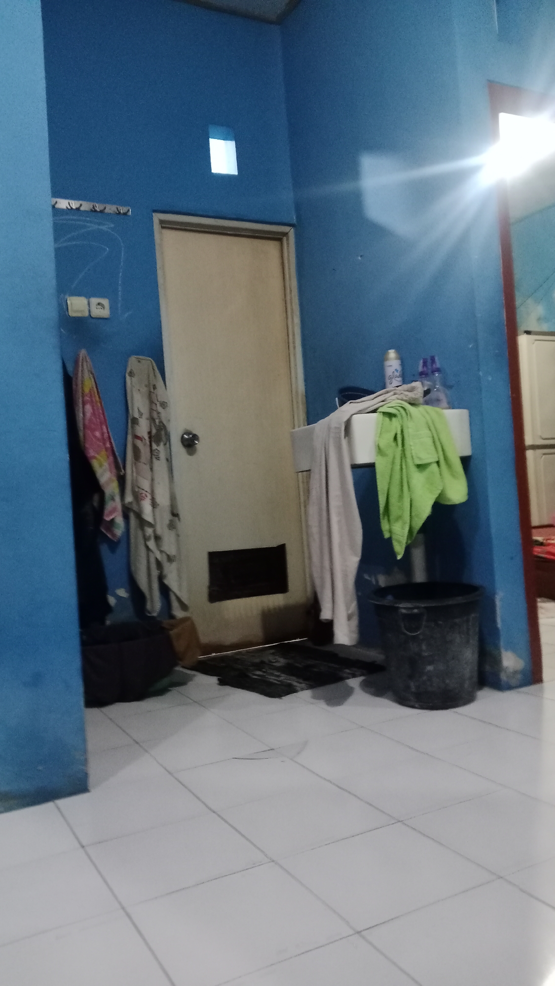 The image depicts an indoor scene, likely a bathroom or utility area, with blue painted walls and white tiled flooring. The room appears somewhat cluttered.
In the foreground, the white tiled floor is visible, with some grout lines present. To the left, a portion of a blue wall juts into the frame. On this wall, there are some faint chalk or marker drawings, and what appear to be two electrical outlets. Two towels are hanging on this wall: one is a multi-colored towel with pink, yellow, and blue patterns, and the other is a cream-colored towel with a subtle pattern. Below these towels, a dark-colored laundry basket or bin is situated.
Dominating the center of the image is a light-colored door with a silver doorknob. To the right of the door, a white sink is mounted on the blue wall. A light-colored towel is draped over the left side of the sink, and a bright green towel is draped over the right side. On the sink counter, there are two spray bottles, one with a white label that appears to say "Glade," and another with a purple label. A dark gray plastic bucket with a handle is placed on the floor to the right of the sink.
Further to the right, a portion of a red-framed doorway is visible, leading to another room where some white furniture, possibly cabinets or drawers, can be seen. Bright light, possibly from a window or artificial source, is creating a lens flare effect in the upper right portion of the image.
There are no people visible in the image. The overall impression is of a functional, somewhat untidy domestic space. Without further context, it is impossible to definitively place this scene in Denpasar, Indonesia, though the style of the tiles and the general appearance are consistent with residential interiors found in Southeast Asia.