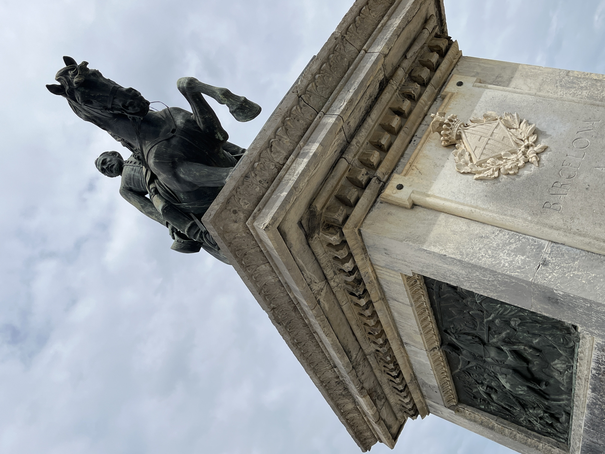 The image depicts a bronze equestrian statue atop a stone pedestal. The statue shows a rider on a rearing horse. The stone pedestal features detailed architectural elements, including decorative moldings, a coat of arms, and the inscription "BARCELONA". The sky is visible in the background, suggesting the statue is outdoors. The location is likely a public square or monument in the city of Barcelona, Spain.