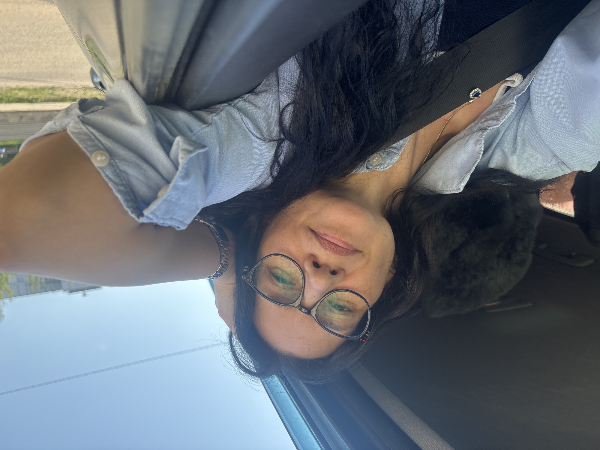 A woman with dark hair and glasses is in the driver's seat of a car, wearing a light blue shirt and a seatbelt. The woman is looking at the camera with a slight smile. The interior of the car is visible. Outside the car window, a glimpse of an urban area is visible, suggestive of Chaikovsky, Russia. The image is rotated such that the sky is oriented towards the left.
