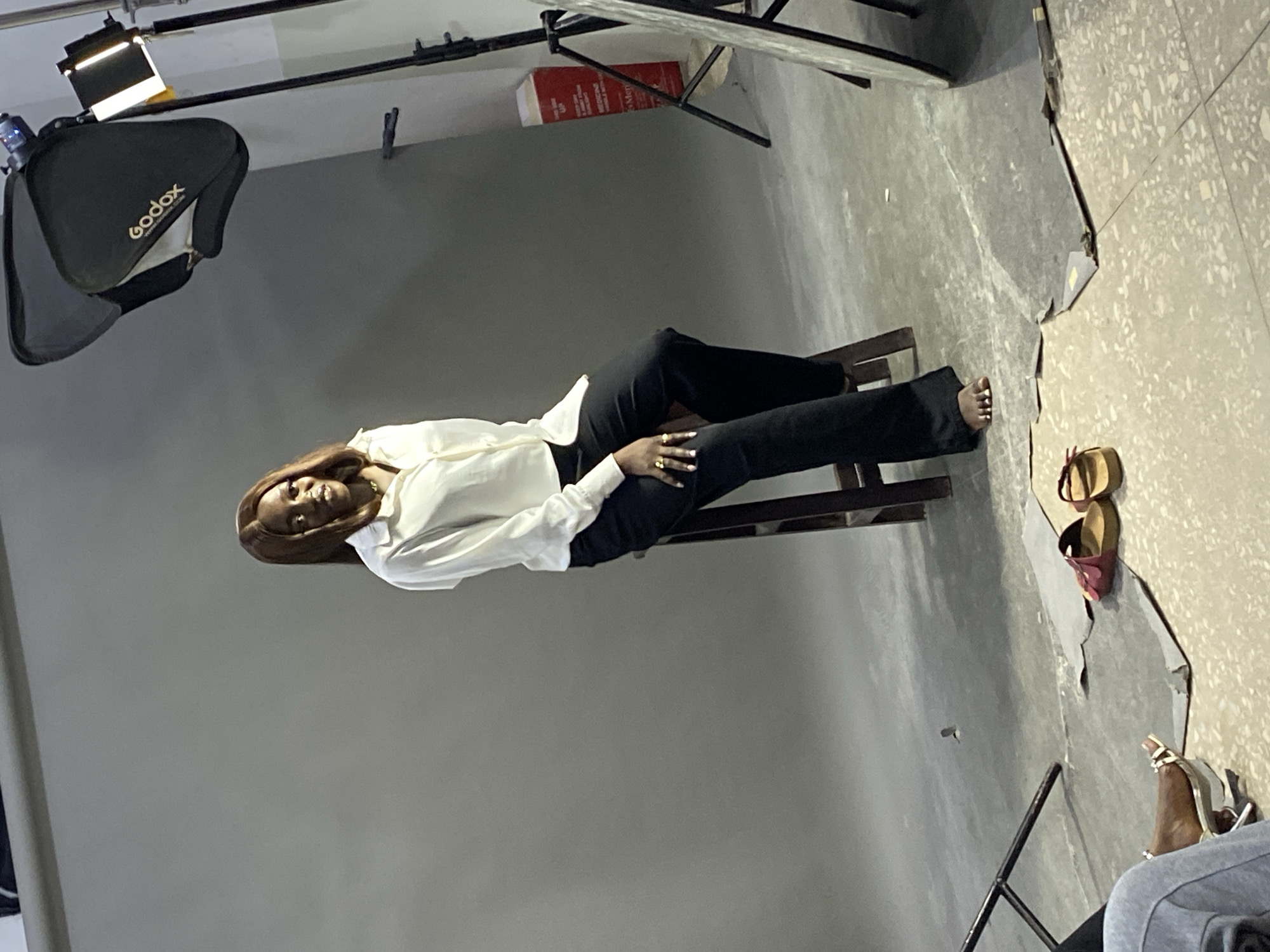 The image shows a woman seated on a wooden stool in a photography studio, likely located in Jos, Nigeria. She is wearing a white button-down shirt and black pants. A "Godox" brand softbox lighting system is positioned above and to the left of her. To the right, on the floor, are several pairs of shoes. A gray backdrop fills most of the frame. Visible studio equipment includes light stands and a red book. The floor is tiled.