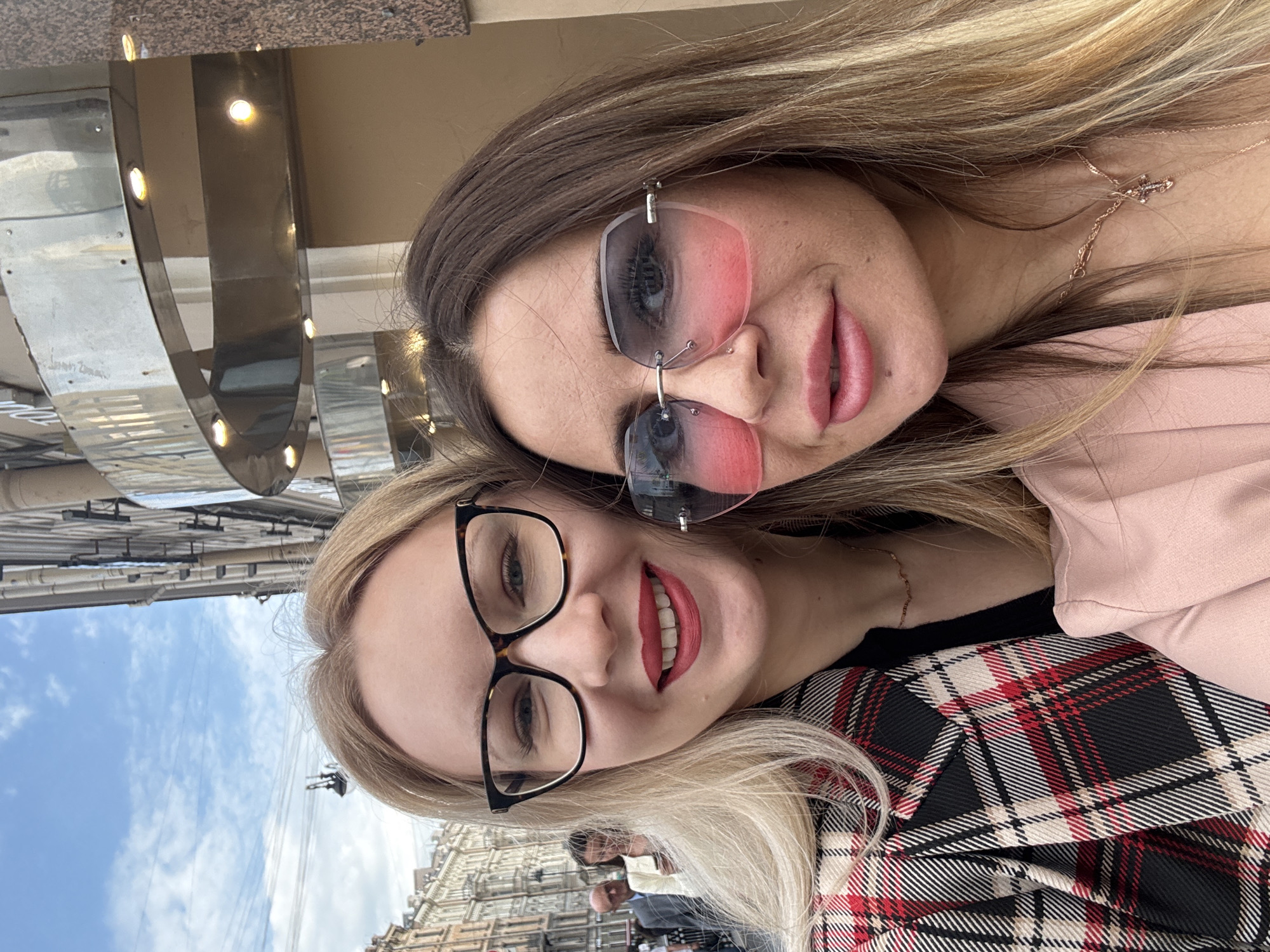 This close-up portrait features two smiling women, posing for a selfie in an urban outdoor setting during the daytime, identified as Saint Petersburg, Russia.
The woman on the right has long blonde hair, blue eyes, and a broad smile, accented by red lipstick. She wears black-rimmed glasses and a black, red, and white plaid jacket over a black top. The woman on the left, with long brown hair highlighted with blonde, offers a subtle smile. She is wearing pink-tinted rimless sunglasses, a light pink top, and a delicate gold-colored necklace with a small cross pendant. A small piercing is visible on her left nostril.
The background, though out of focus, reveals a bright, partly cloudy sky and the facades of architectural buildings, consistent with an older European city. To the upper left, a metallic, reflective structure with embedded circular lights is visible, possibly part of a building's signage or ornamentation. Faint, illegible writing or scratches can be observed on this metallic surface. The overall mood conveyed is cheerful and friendly.