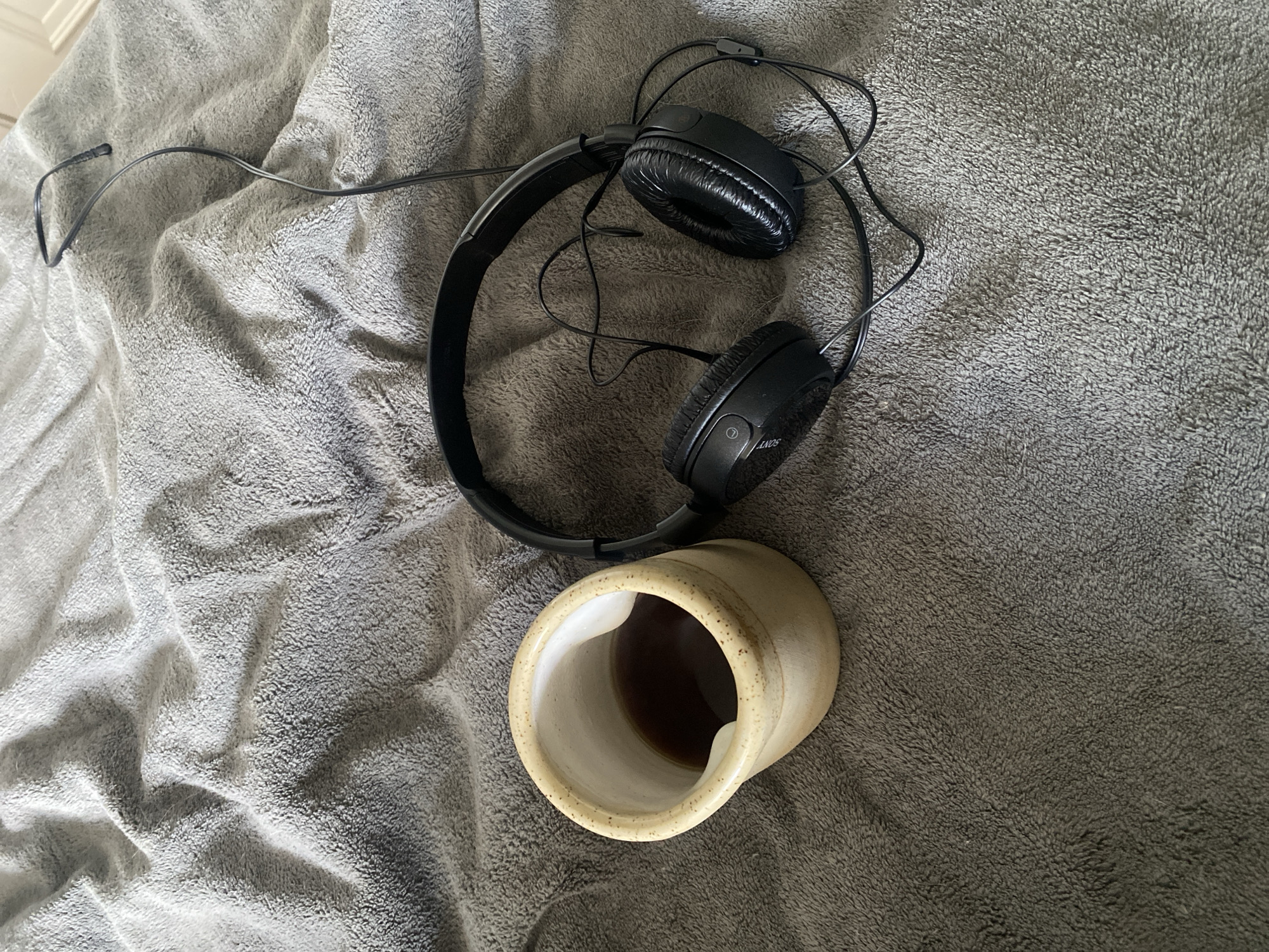 A black Sony headphone set and a ceramic mug are lying on a gray blanket. The mug is tan and speckled, with a white interior and contains an amber liquid. The headphones have a black cord and are positioned partially on top of the mug. The blanket is textured. The location of the scene is not indicated.