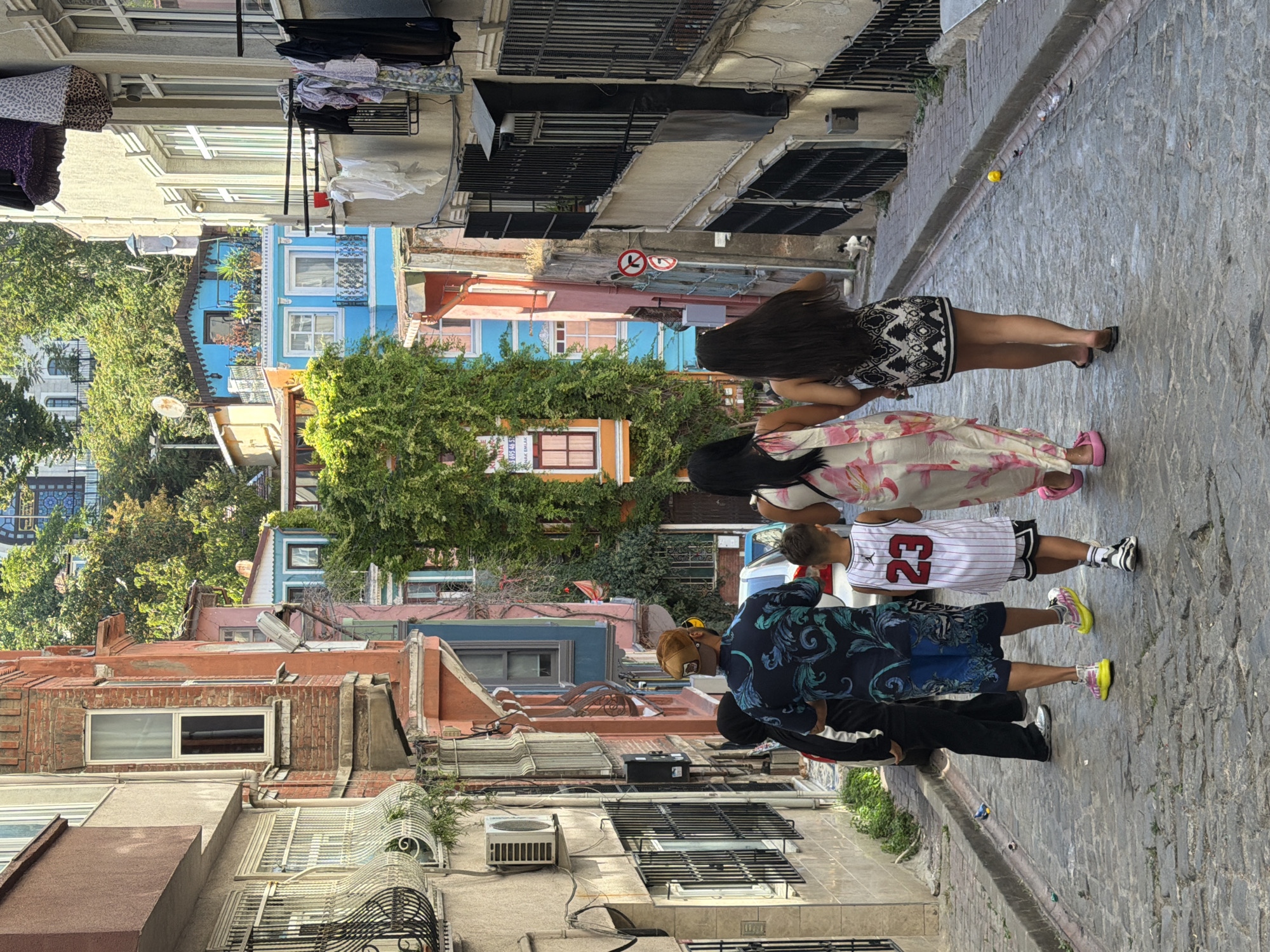 A group of five people walk down a cobblestone street in Istanbul, Turkey. The street is lined with colorful, multi-story buildings. The people are walking away from the camera and appear to be a family. From left to right, there is a person in a black hooded garment, a person in a dark blue patterned shirt and blue shorts, a young boy in a white jersey with the number 23, a woman in a long, floral dress, and a woman in a short, black and white patterned dress. In the background, a lush green tree dominates the scene, with buildings of various colors, including blue, pink, and orange, peeking through the foliage. The architecture suggests a historic district. The sun is shining, casting shadows on the street.