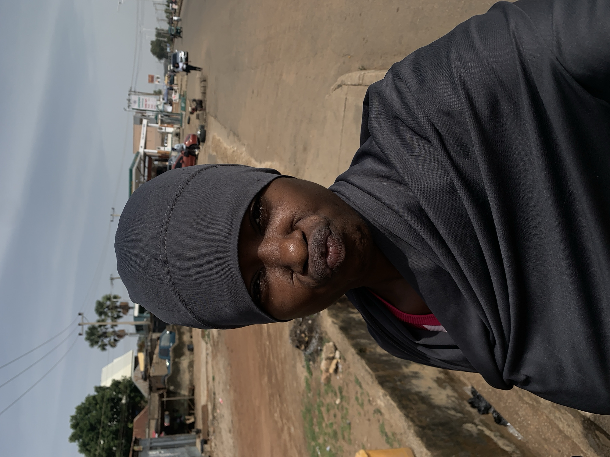 A medium, eye-level shot depicts a woman in Rafin Par, Nigeria, wearing a gray head covering and matching robe. She is standing on a street with a cracked pavement and an open drain adjacent to the road. Litter and debris are visible in the drain. In the background, on the opposite side of the street, are buildings, trees, and power lines, with several vehicles and people scattered along the road. The sky is overcast. The woman has dark skin and pursed lips.