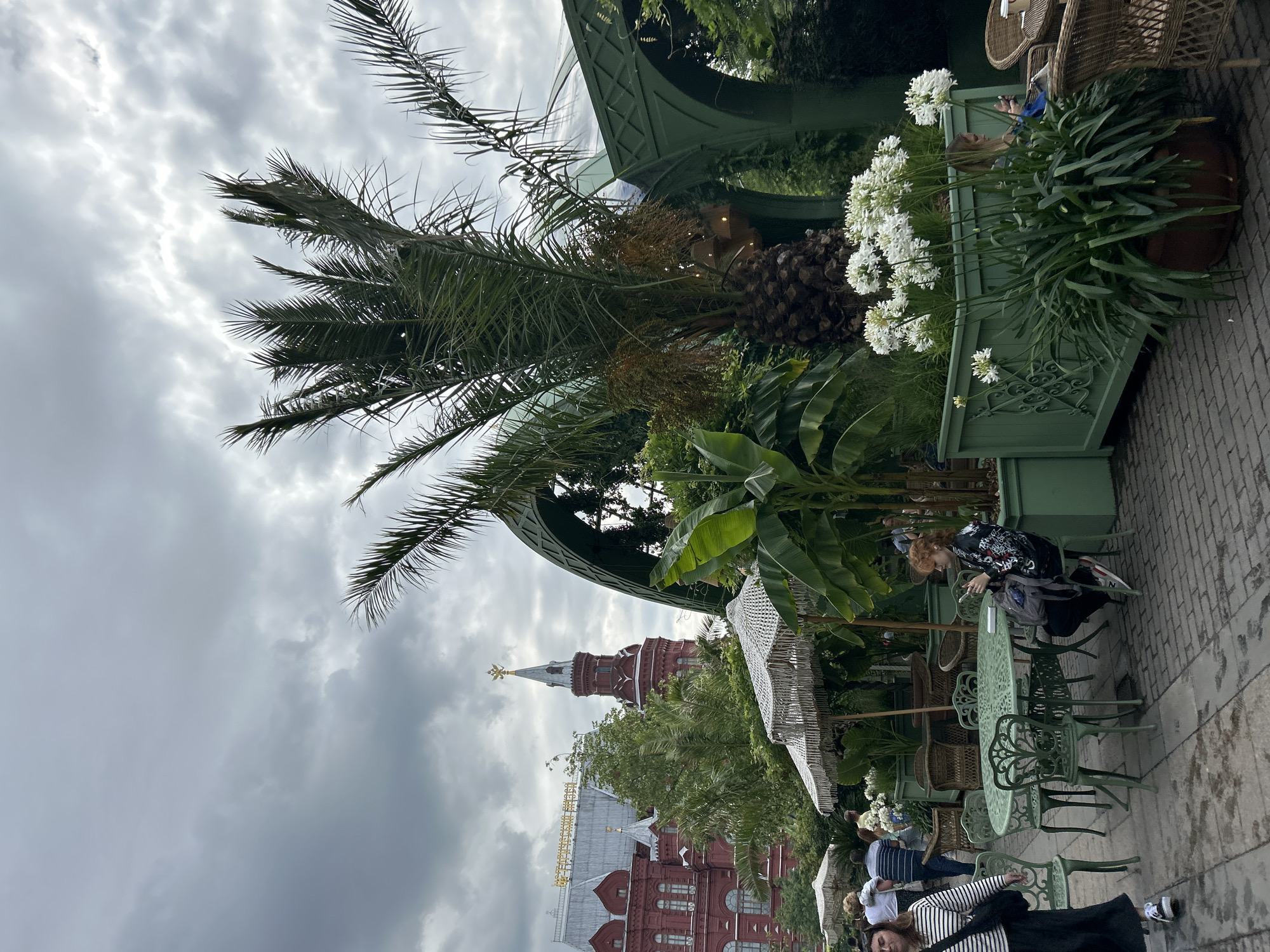 The image depicts an outdoor seating area with lush greenery and a palm tree, set against the backdrop of a historic red building with a distinct tower, identified as the State Historical Museum in Moscow, Russia. In the foreground, a woman with red hair is seated at a green metal table, looking at her phone. To her left, a woman in a striped shirt walks past. Several other people are visible in the background, some walking and others seated at tables. The environment suggests a restaurant or cafe setting, with green metal tables and chairs, umbrellas, and potted plants. The sky is overcast, indicating a cloudy day.
