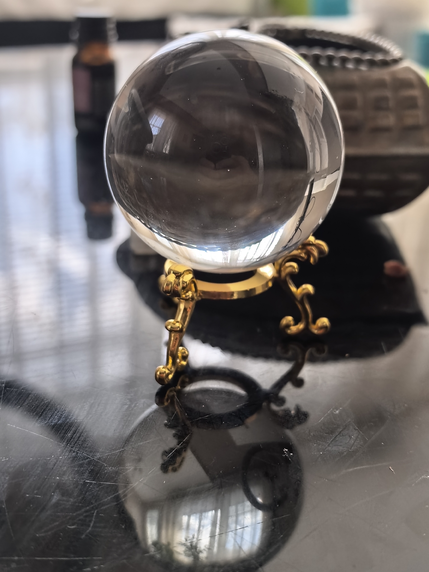 On a reflective surface, likely a tabletop, rests a clear crystal ball held by a golden ornamental stand. To the left and behind the ball is a small, dark-colored bottle, and to the right is a larger, dark, textured container. The surface shows reflections of nearby objects, including a window, revealing an exterior scene of Unknown, Unknown.